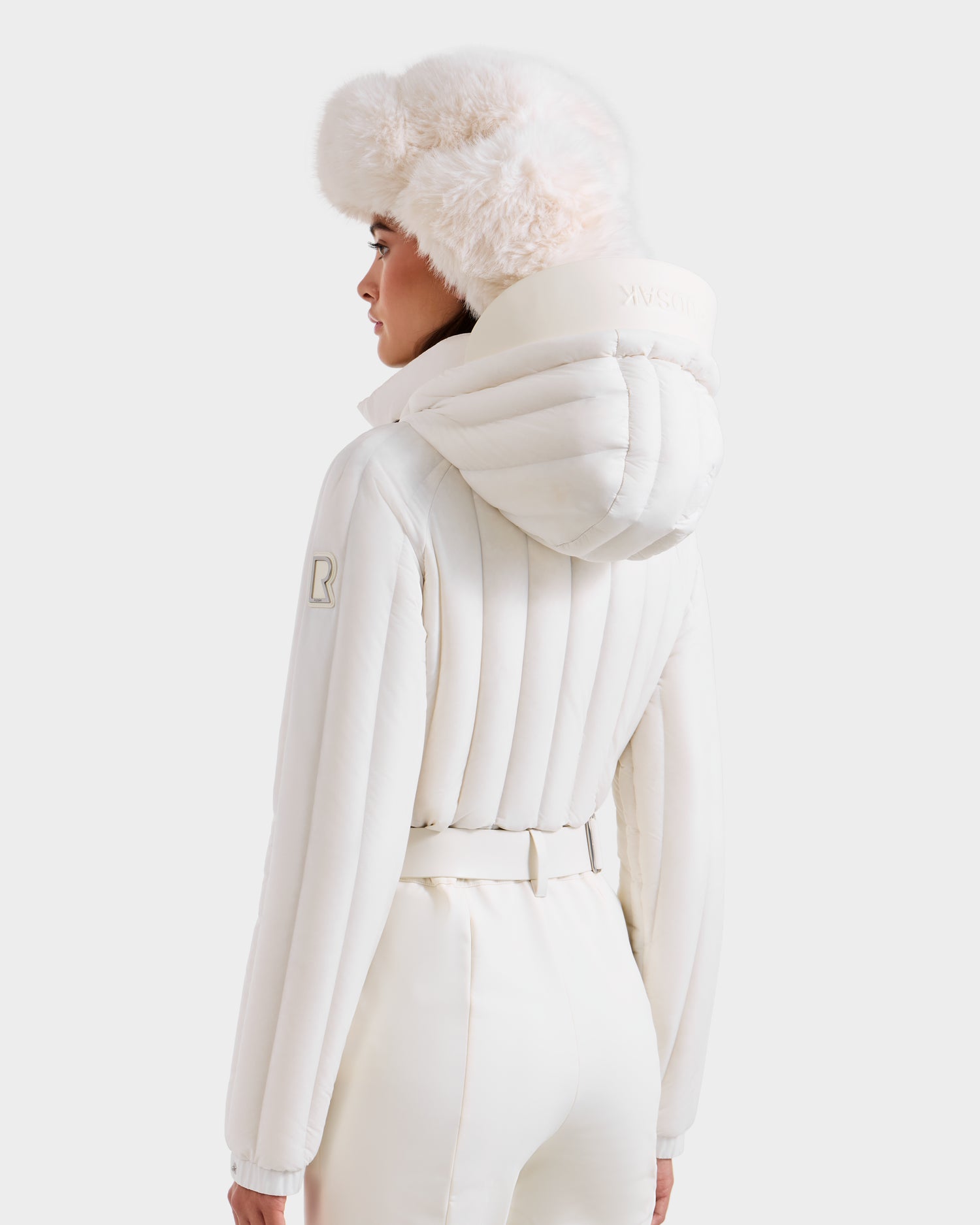 A person wearing the Rudsak SAGE - 8124782 Iceland all-white padded winter jacket with a matching belt and high collar, plus a fluffy white fur hat, stands against a plain white background, facing away from the camera.