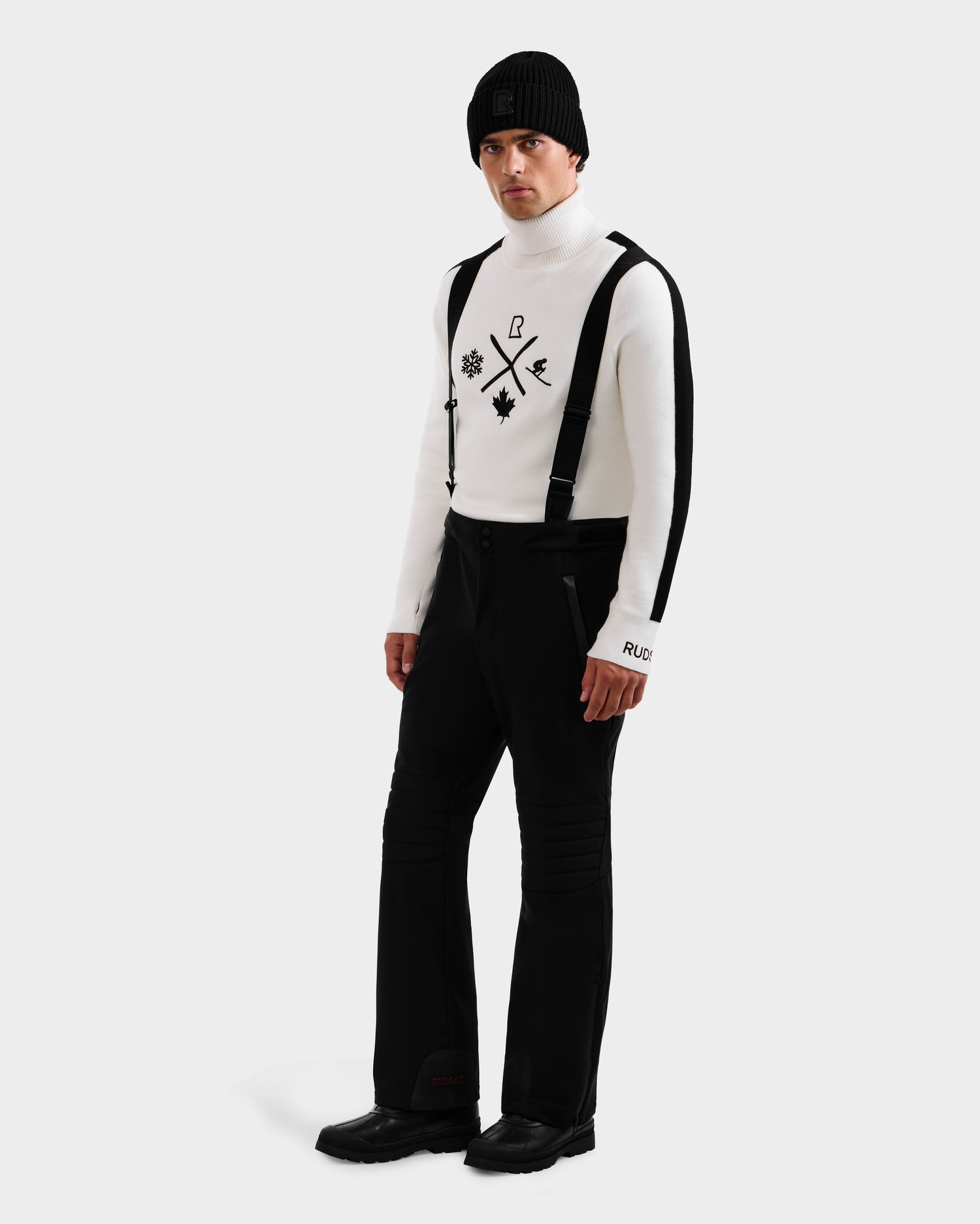 A man wears the Rudsak SAMPSON - 6125586 Black sweater with black graphics, paired with black suspenders, pants, boots, and a beanie, standing against a plain light background.