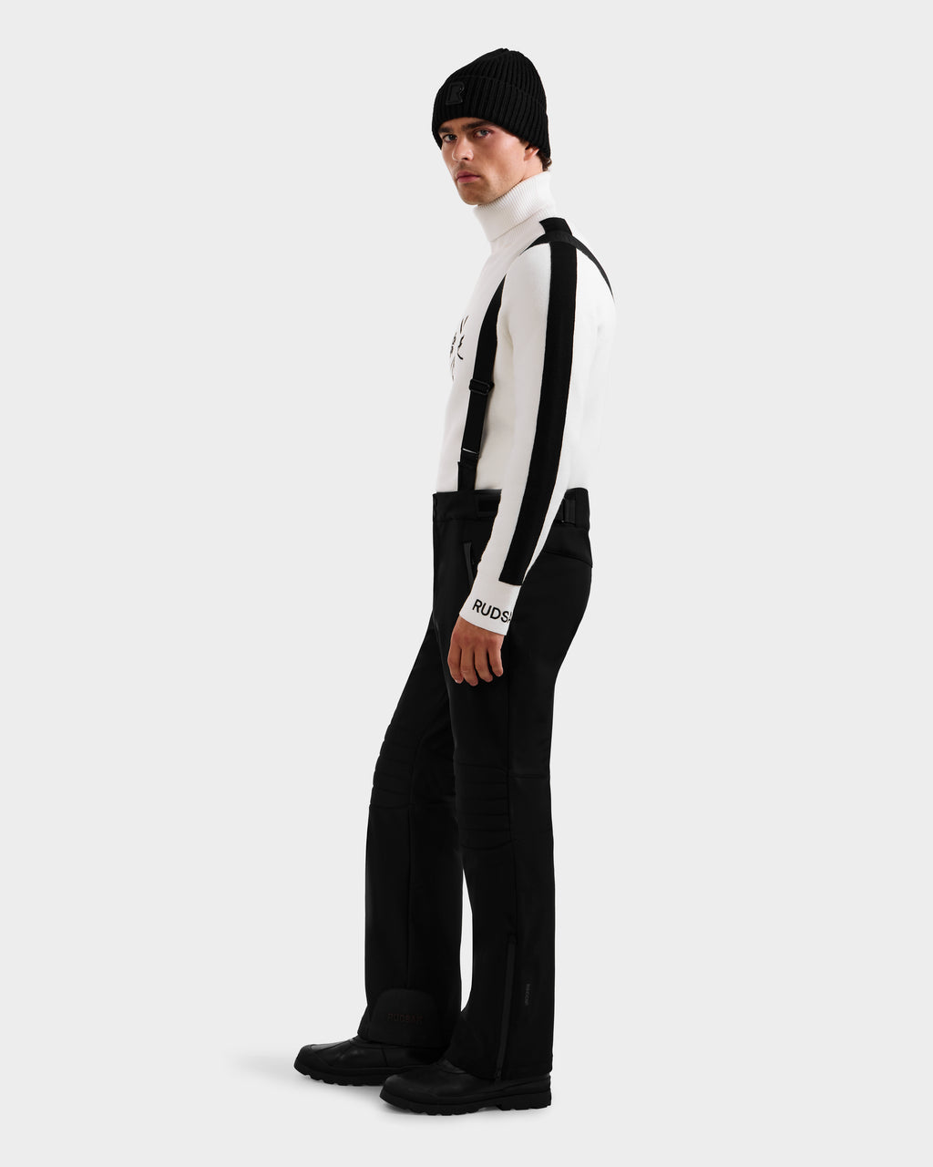 Wearing the Rudsak SAMPSON - 6125586 Black beanie, a person in a white striped turtleneck, black suspenders, pants, and boots stands against a plain light gray background, looking slightly over their shoulder.