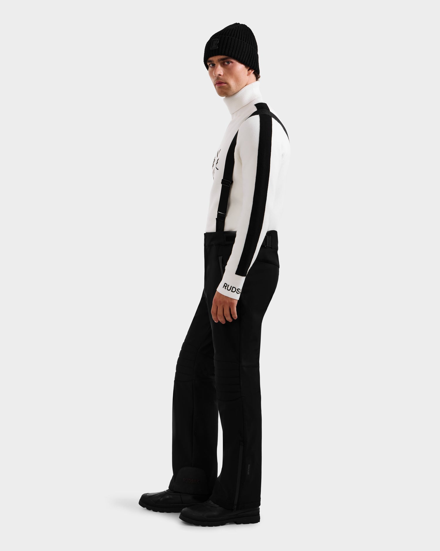 Wearing the Rudsak SAMPSON - 6125586 Black beanie, a person in a white striped turtleneck, black suspenders, pants, and boots stands against a plain light gray background, looking slightly over their shoulder.