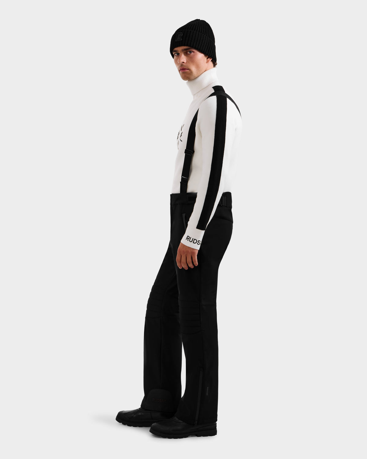Wearing the Rudsak SAMPSON - 6125586 Black beanie, a person in a white striped turtleneck, black suspenders, pants, and boots stands against a plain light gray background, looking slightly over their shoulder.