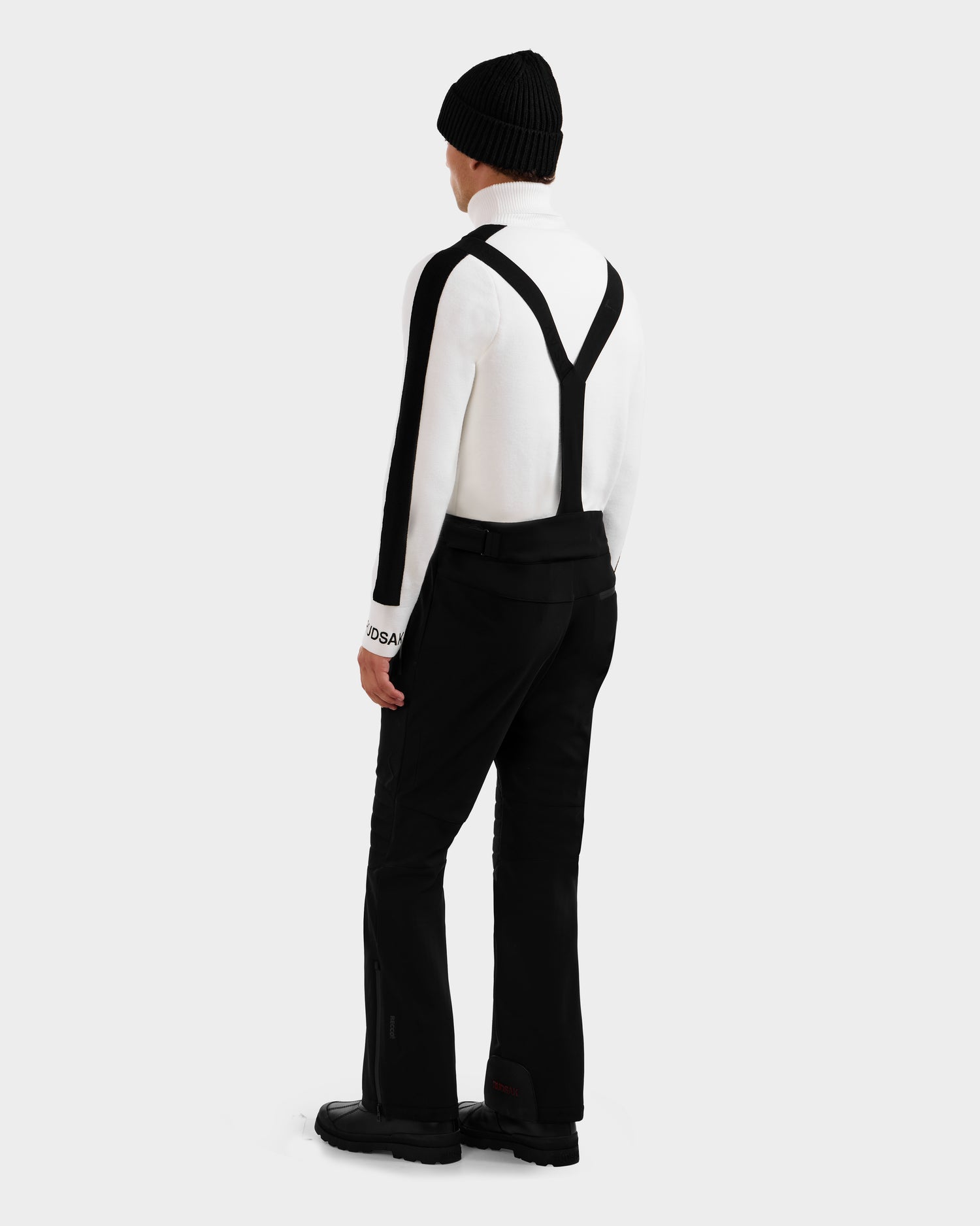 A person wearing the Rudsak SAMPSON - 6125586 Black stands facing away against a plain light gray background, styled with a white long-sleeve top, black suspenders, black pants, black boots, and a black beanie.