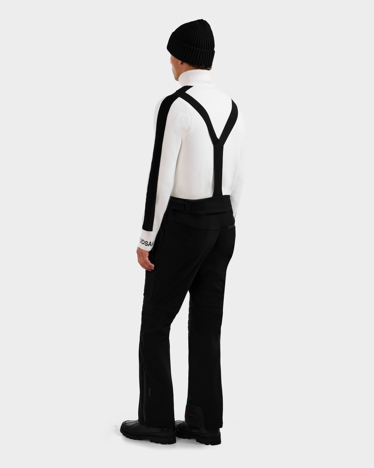 A person wearing the Rudsak SAMPSON - 6125586 Black stands facing away against a plain light gray background, styled with a white long-sleeve top, black suspenders, black pants, black boots, and a black beanie.