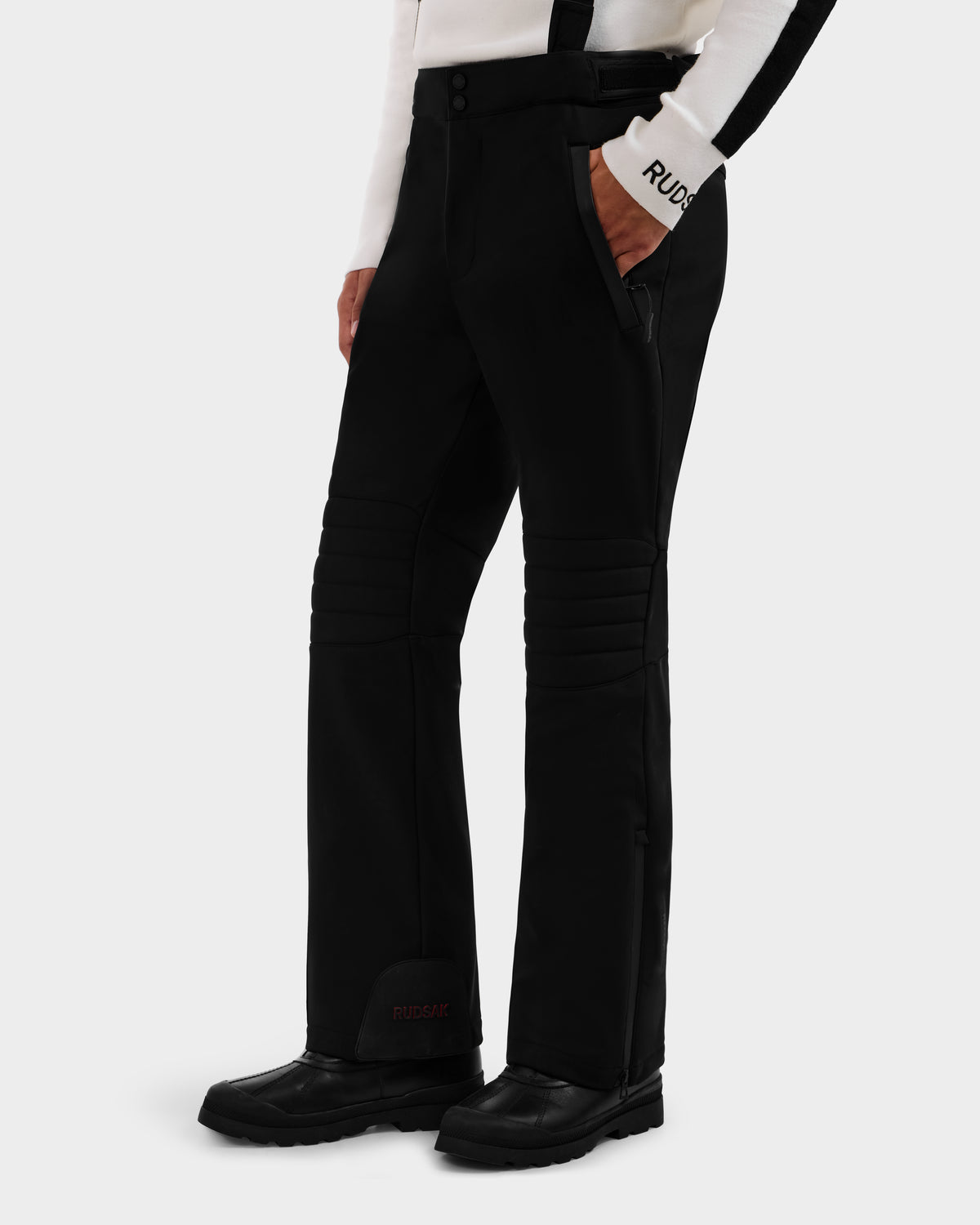 A person wears the Rudsak SAMPSON - 6125586 Black men’s performance snow pant with waterproof zippers, black boots, and a white and black long-sleeve top featuring RUDSAK text on the sleeve, posing hands-in-pockets against a plain background.
