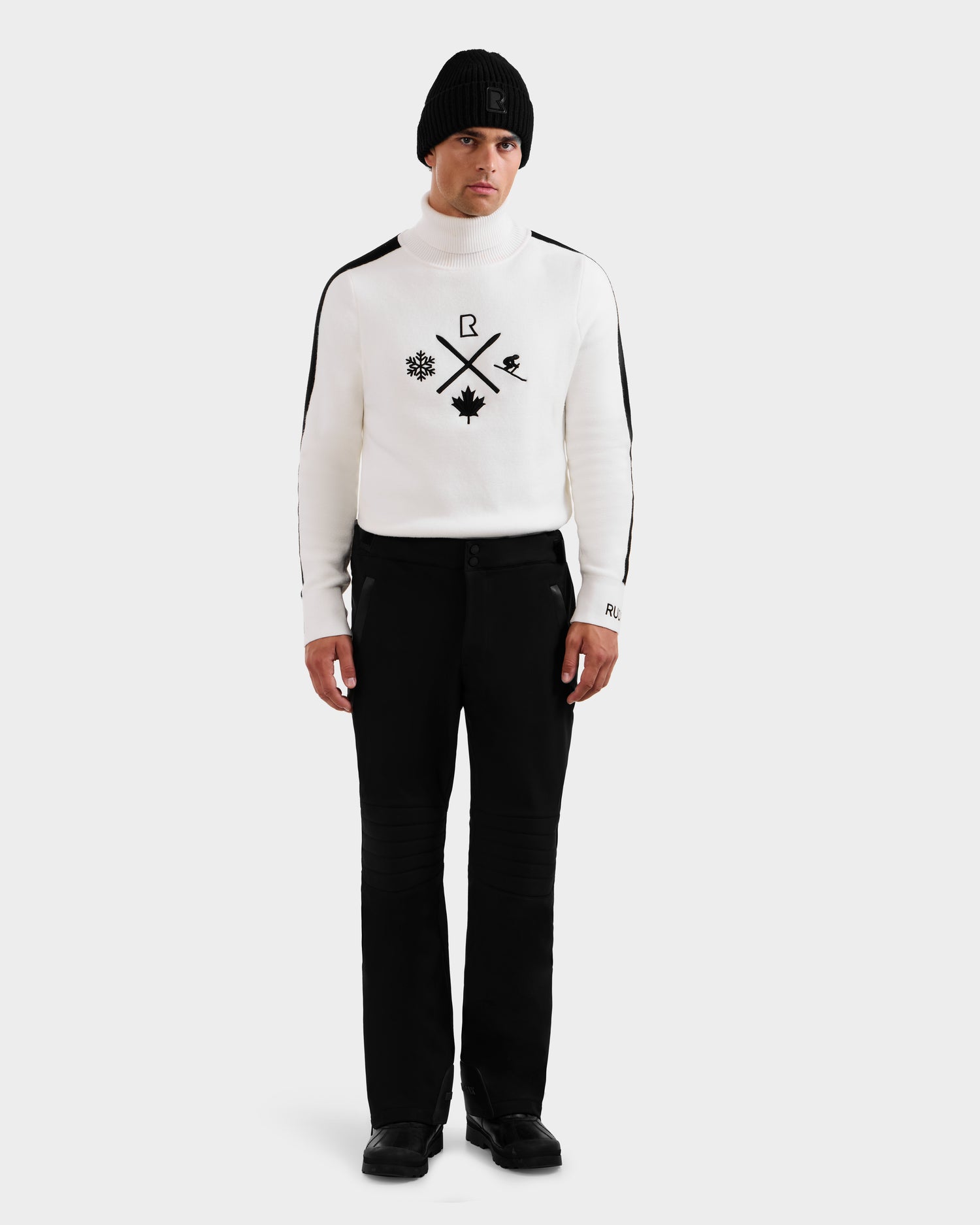 A man models the Rudsak SAMPSON - 6125586 Black beanie and a white turtleneck sweater with black symbols, paired with black pants and shoes, standing against a plain white background.