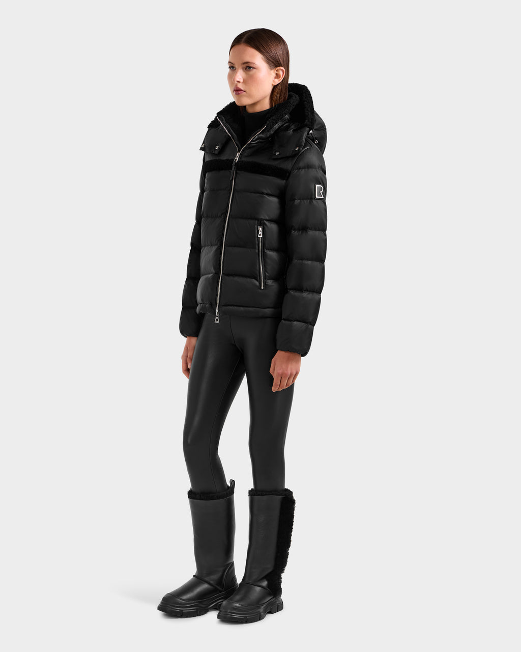 A woman with straight brown hair and a neutral expression models the Rudsak SANDRA - 8125612 Black women’s shearling puffer jacket, paired with black shiny leggings and fur-trimmed boots, showcasing contemporary style and lightweight insulation.