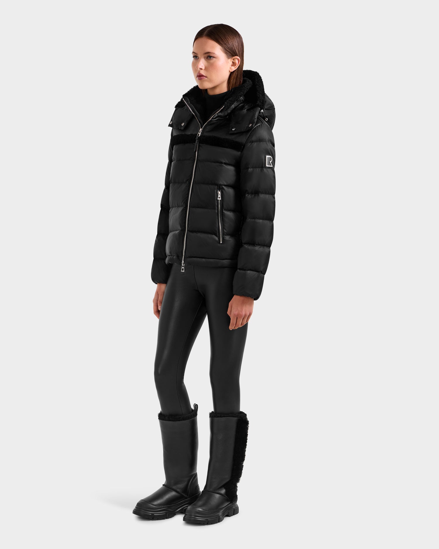 A woman with straight brown hair and a neutral expression models the Rudsak SANDRA - 8125612 Black women’s shearling puffer jacket, paired with black shiny leggings and fur-trimmed boots, showcasing contemporary style and lightweight insulation.
