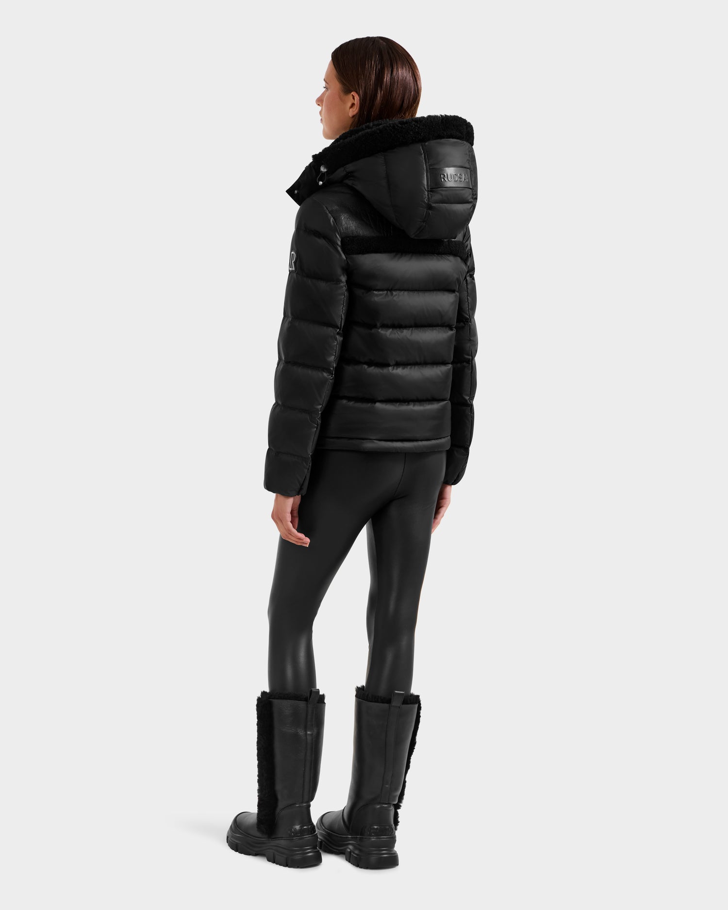 A person with long brown hair stands facing away, wearing the Rudsak SANDRA - 8125612 Black women’s shearling puffer jacket with a hood, shiny black leggings, and tall black boots with fur trim against a light gray background.