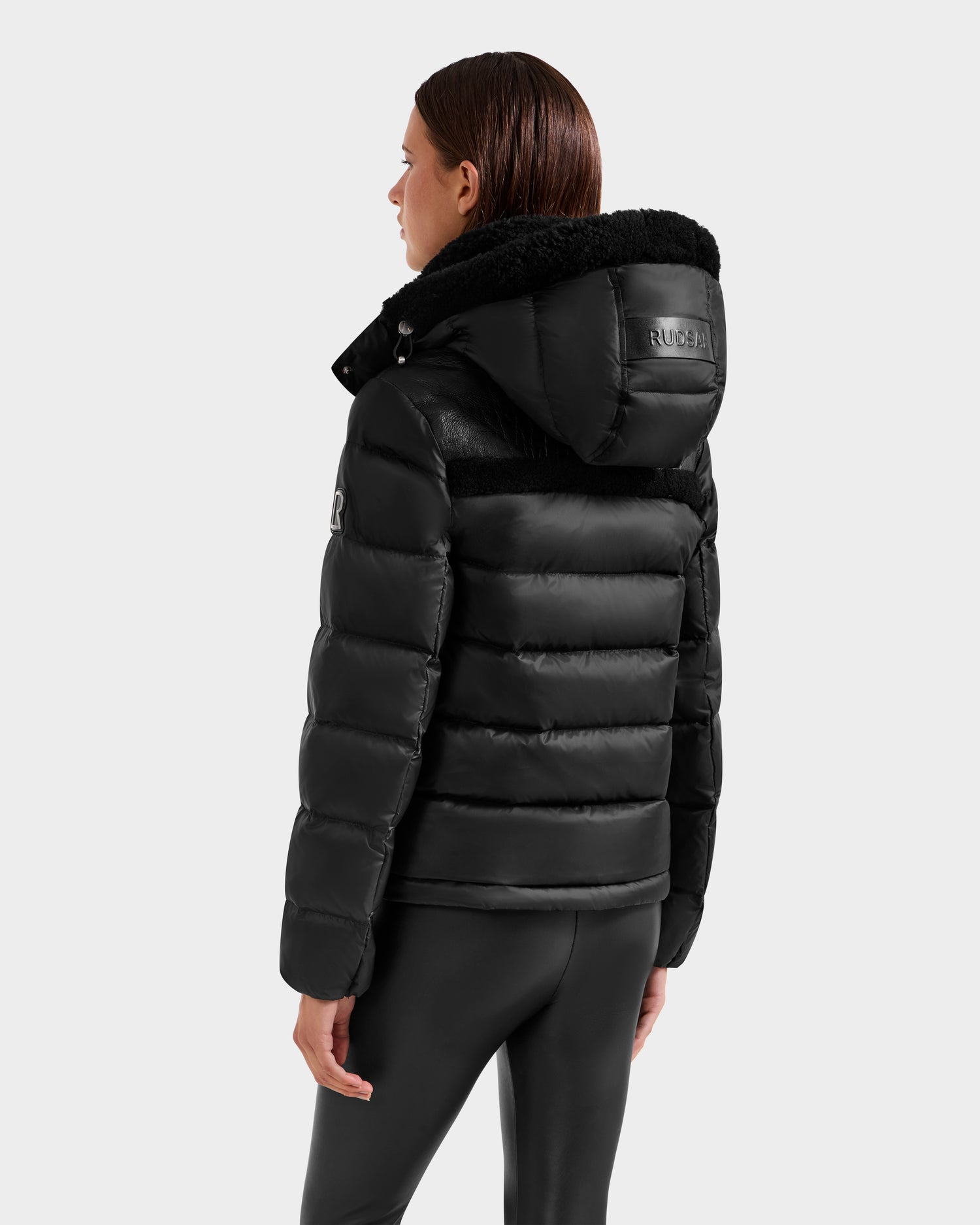 A person wearing the Rudsak SANDRA - 8125612 Black puffer jacket and black leggings stands facing away, displaying the jacket's shiny finish and patches on the back and left arm. The contemporary style features lightweight insulation.