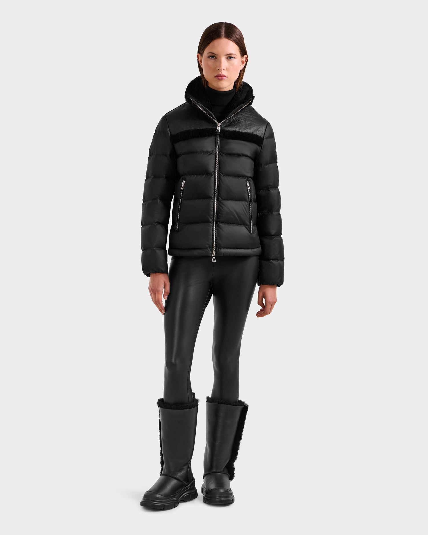 A woman faces forward in a contemporary style, wearing the Rudsak SANDRA - 8125612 Black shearling puffer jacket with black faux leather leggings and tall fur-lined winter boots, set against a plain light gray background.
