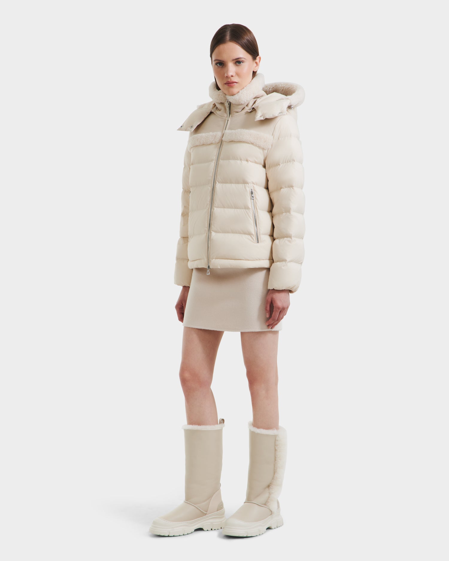 A woman poses against a white background wearing the Rudsak SANDRA - 8125612 Vanilla shearling puffer jacket with lightweight insulation, paired with a matching knee-length skirt and tall beige winter boots for a cozy, modern style.