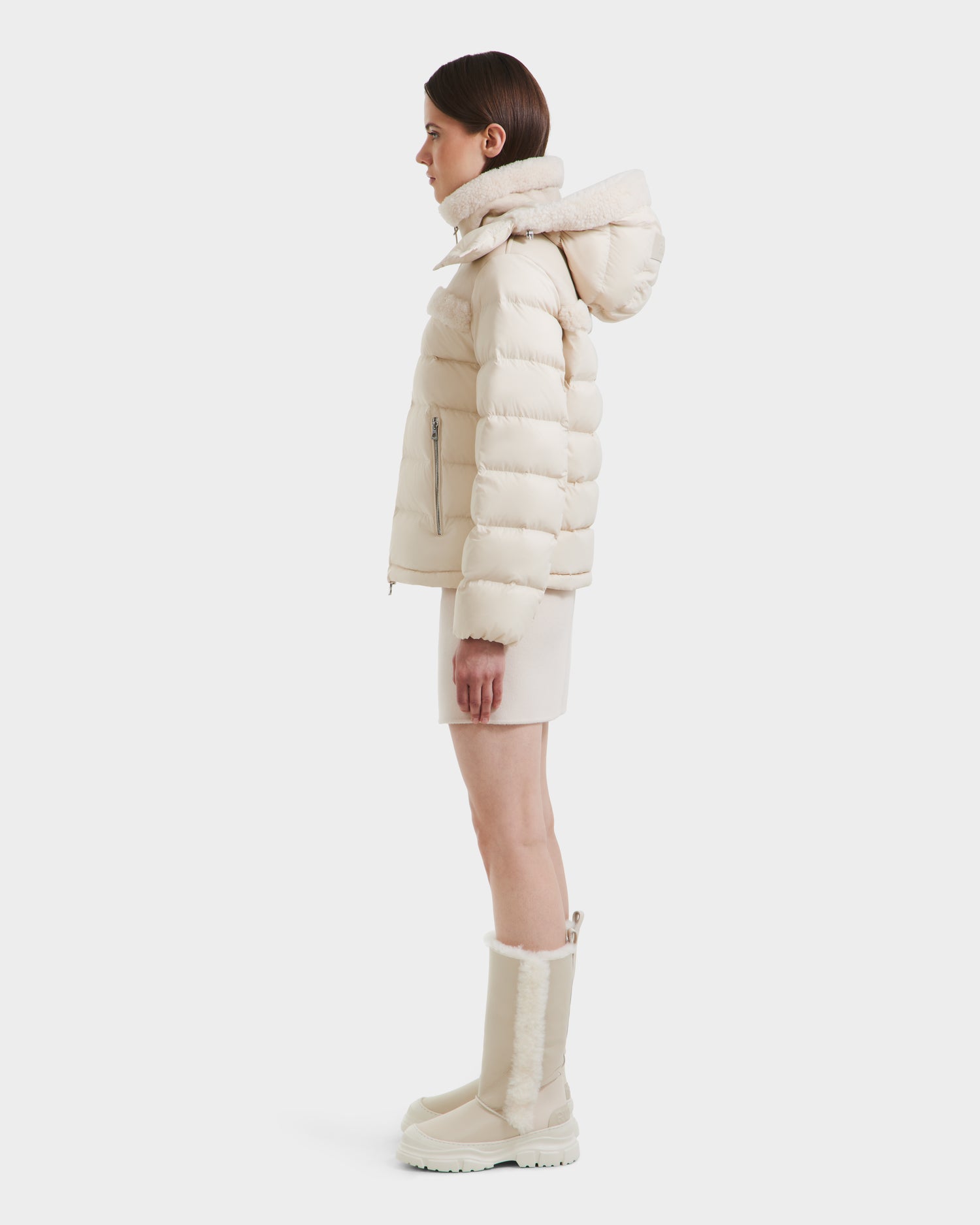 A woman stands in profile against a plain white background wearing the Rudsak SANDRA - 8125612 Vanilla women's shearling puffer jacket with lightweight insulation, a matching skirt, and tall fur-lined boots.