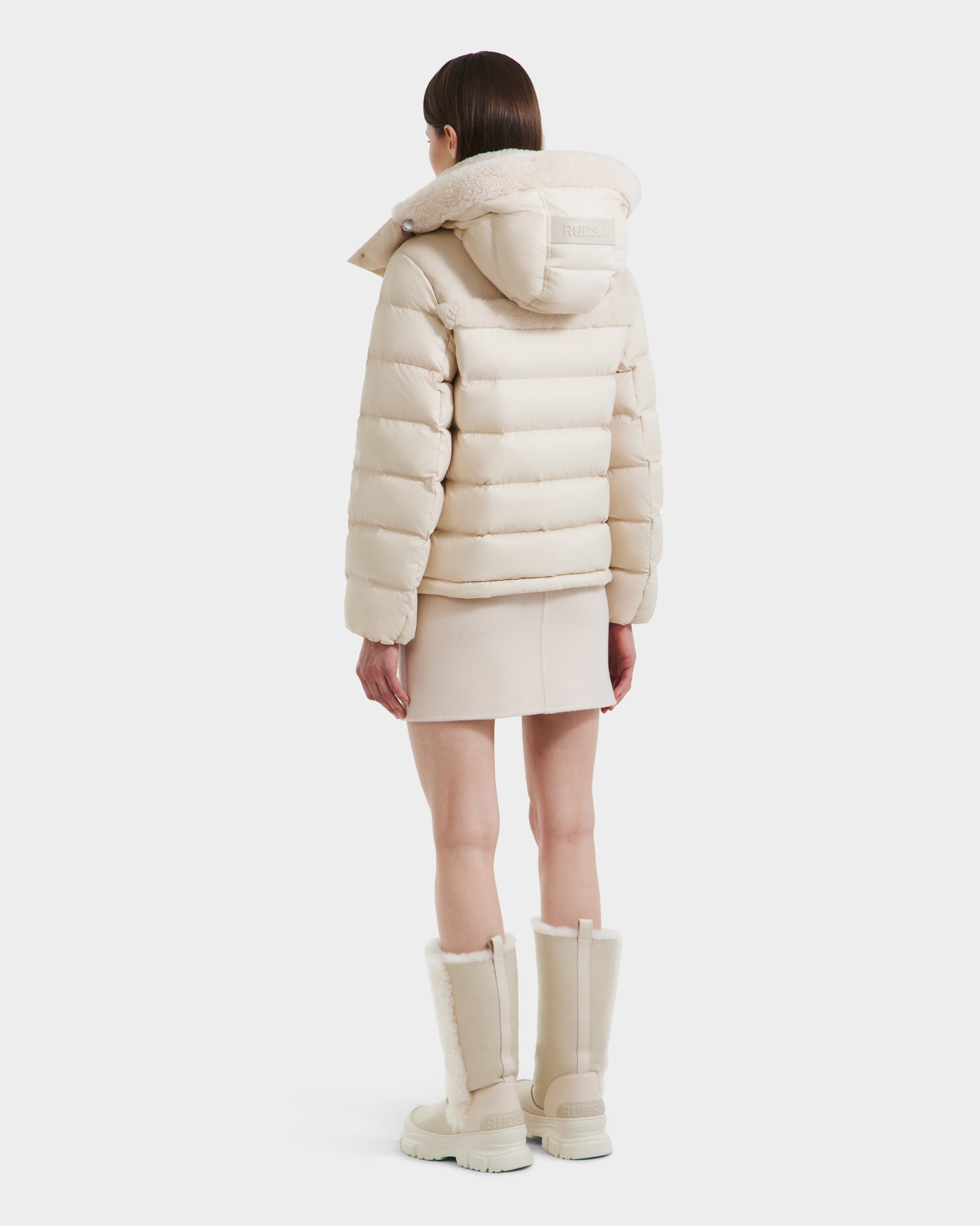 A woman faces away, modeling the Rudsak SANDRA - 8125612 Vanilla women's shearling puffer jacket with lightweight insulation, styled with a matching short skirt and fur-trimmed knee-high boots in light beige against a white background.