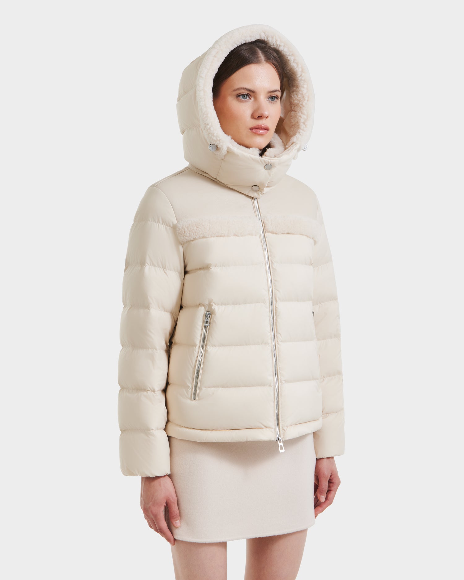 A woman wears the Rudsak SANDRA - 8125612 Vanilla shearling puffer jacket with faux fur hood, made from recycled nylon and down-feather fill, paired with a matching beige skirt against a plain white background.