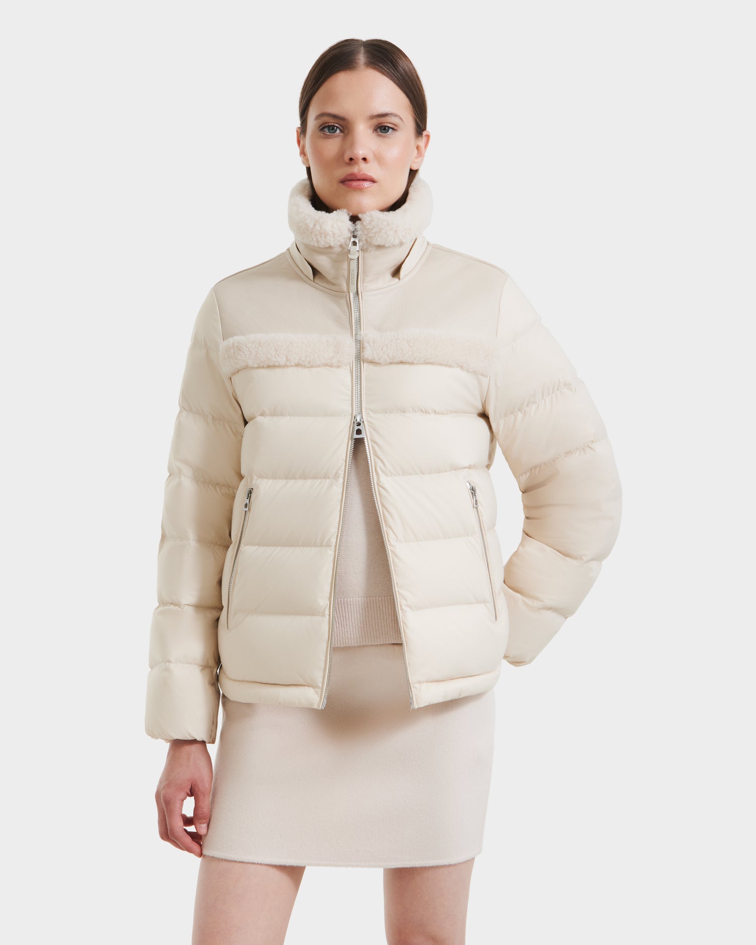 A woman with straight brown hair stands with one hand in her pocket, wearing the Rudsak SANDRA - 8125612 Vanilla shearling puffer jacket. She styles its high collar with a matching skirt and top for a modern, coordinated look.