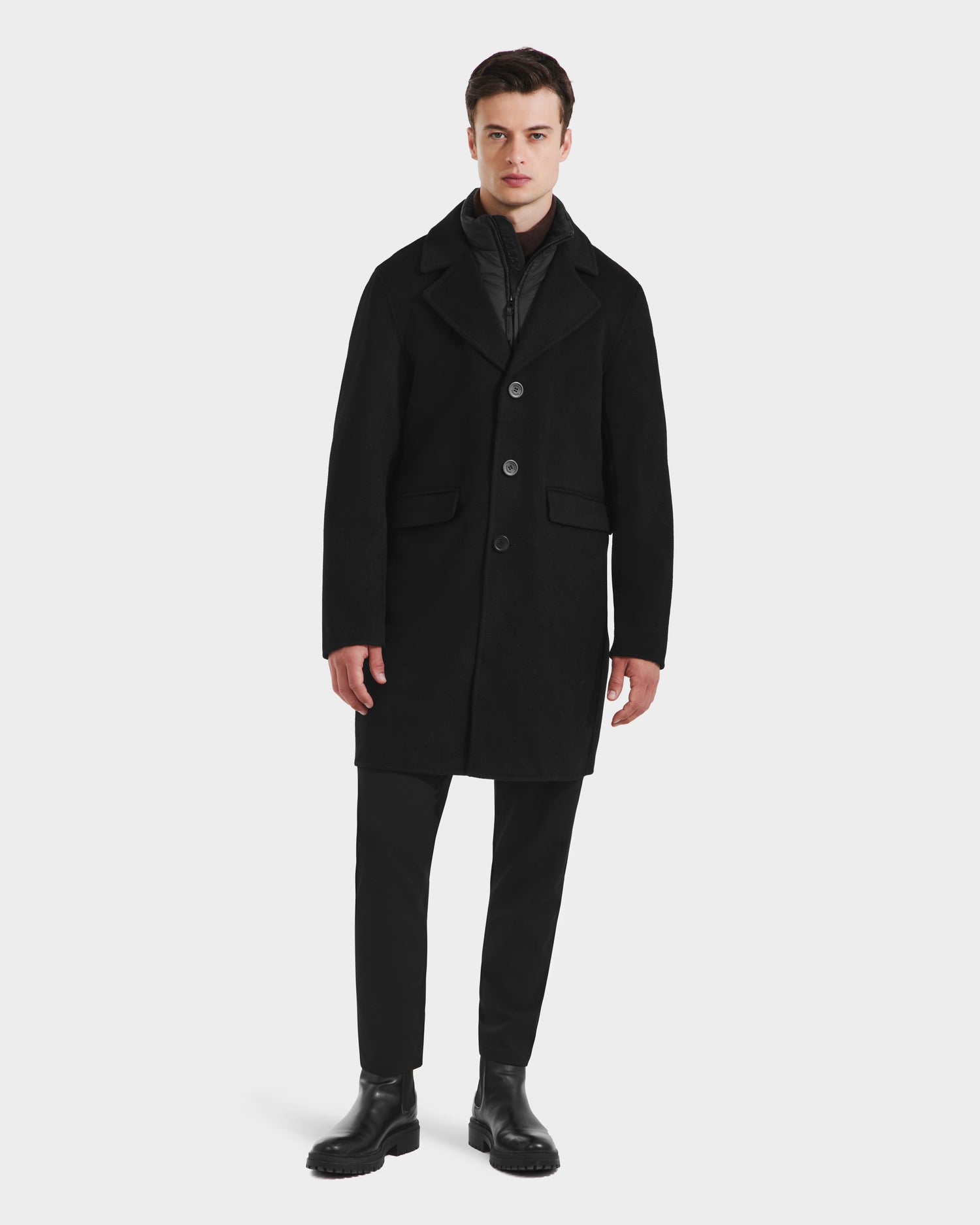 A man stands against a plain white background wearing the Rudsak SAWYER - 6125511 Black men’s wool blend coat, paired with black pants, black boots, and a dark inner layer. He looks straight ahead with a neutral expression and relaxed posture.