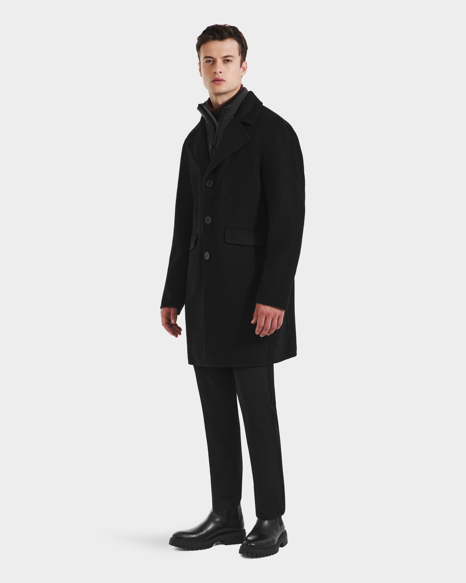 A man faces slightly to the side against a white background, wearing the Rudsak SAWYER - 6125511 Black men’s wool blend coat, paired with black trousers, boots, and a dark zip-up top.