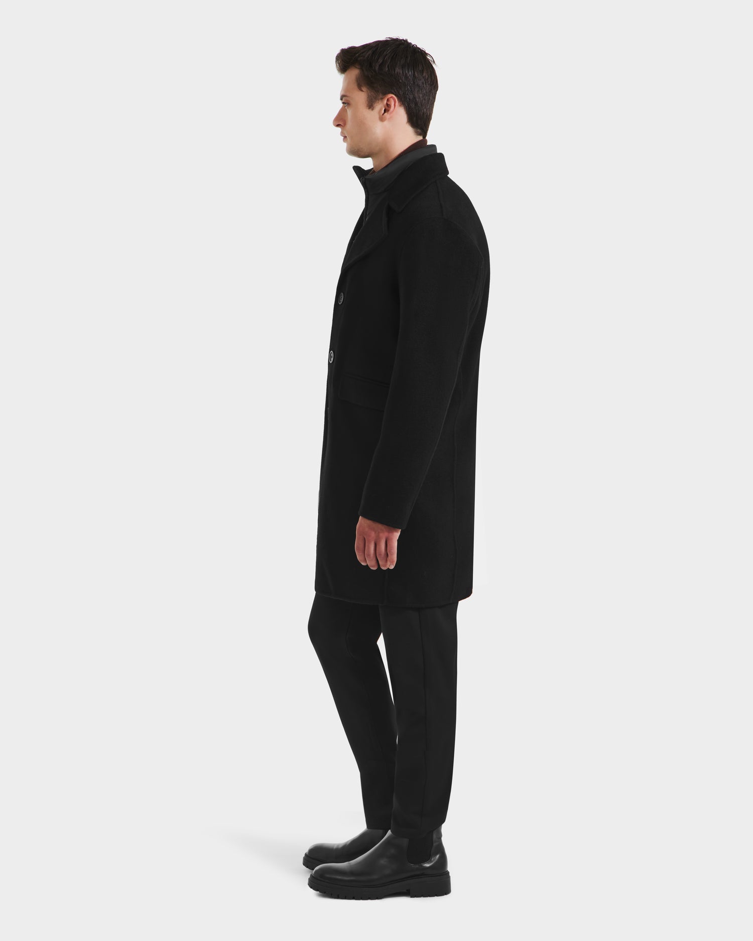 A man stands in profile facing left, wearing the Rudsak SAWYER - 6125511 Black wool blend coat with a removable quilted dickie, paired with black pants and boots against a plain white background.