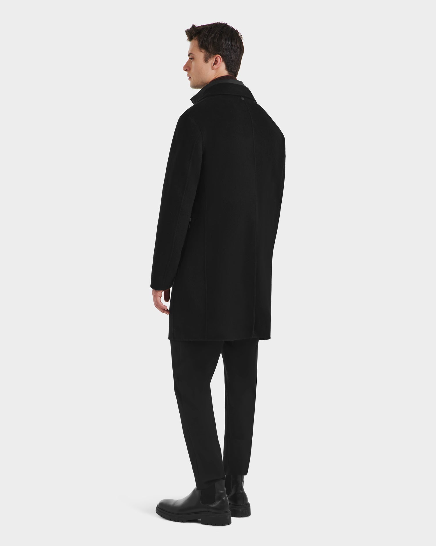 A man stands facing away, wearing the Rudsak SAWYER - 6125511 Black mens wool blend coat with a classic silhouette, paired with black pants and boots against a plain light background.