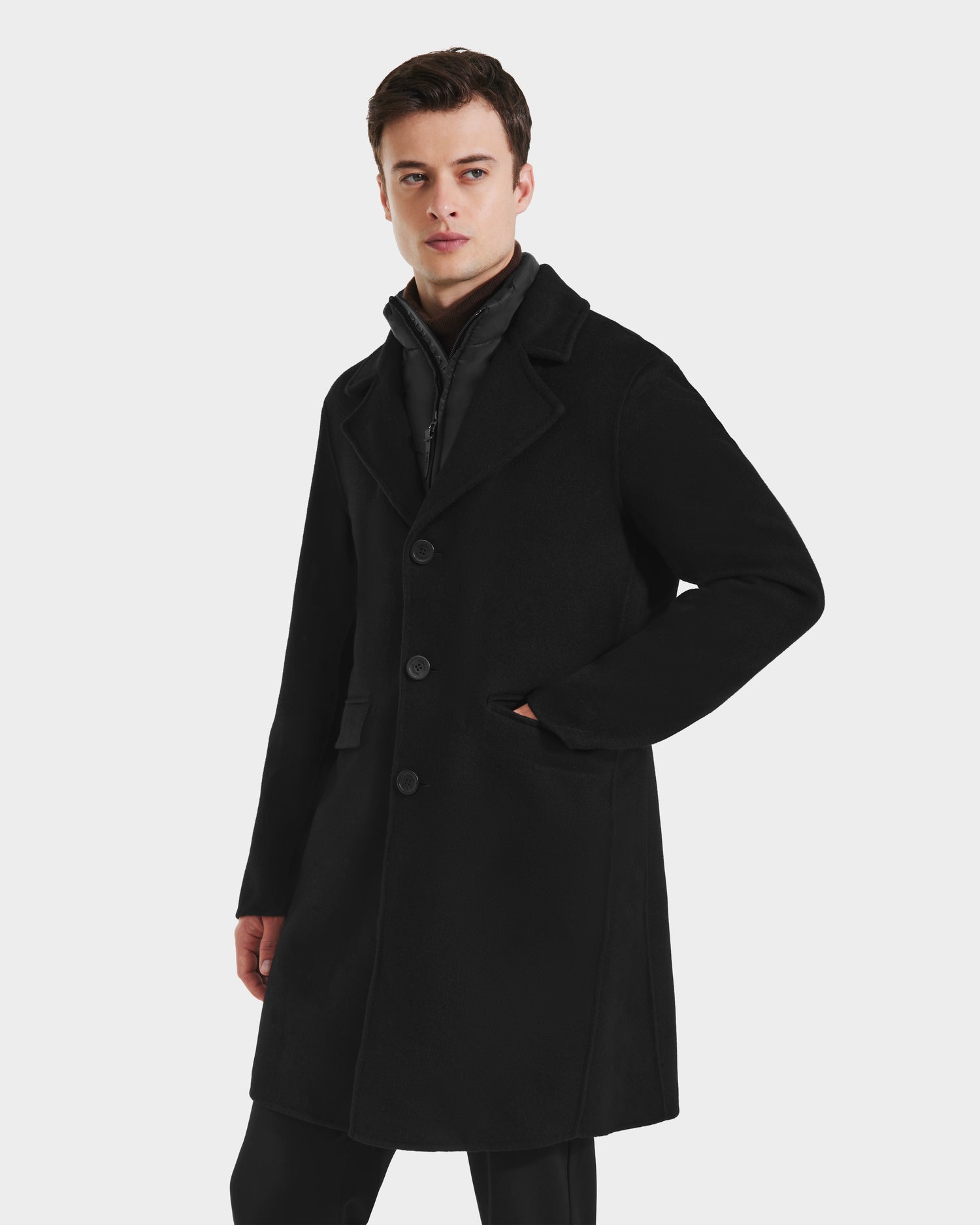 A young man wearing the Rudsak SAWYER - 6125511 Black wool blend coat with a classic silhouette stands against a plain white background, one hand in his pocket and a neutral expression.