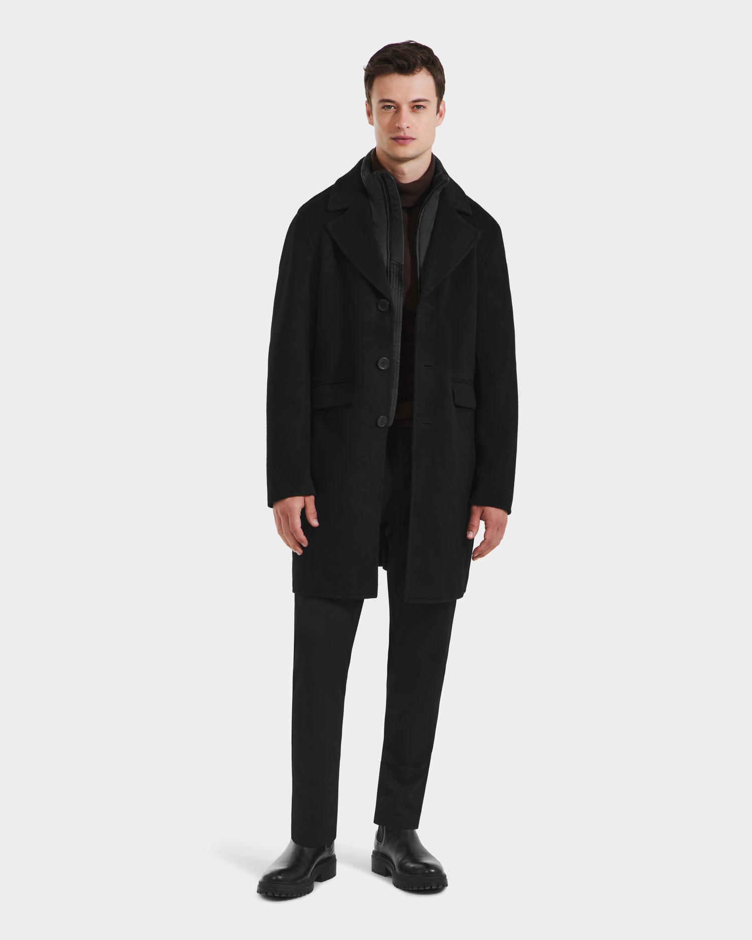 A man faces forward in a classic silhouette, wearing the Rudsak SAWYER - 6125511 Black men’s wool blend coat with black pants, black shoes, and a dark scarf, standing against a plain white background.