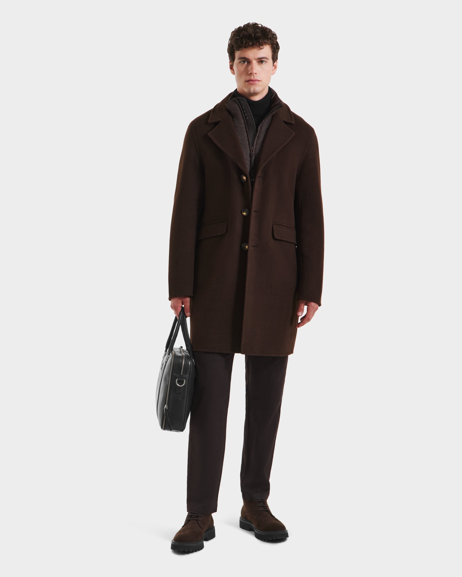 A man stands facing forward against a plain light background, wearing the Rudsak SAWYER - 6125511 Espresso dark brown wool blend coat, paired with dark pants, dress shoes, and holding a black leather briefcase. He has short curly hair.