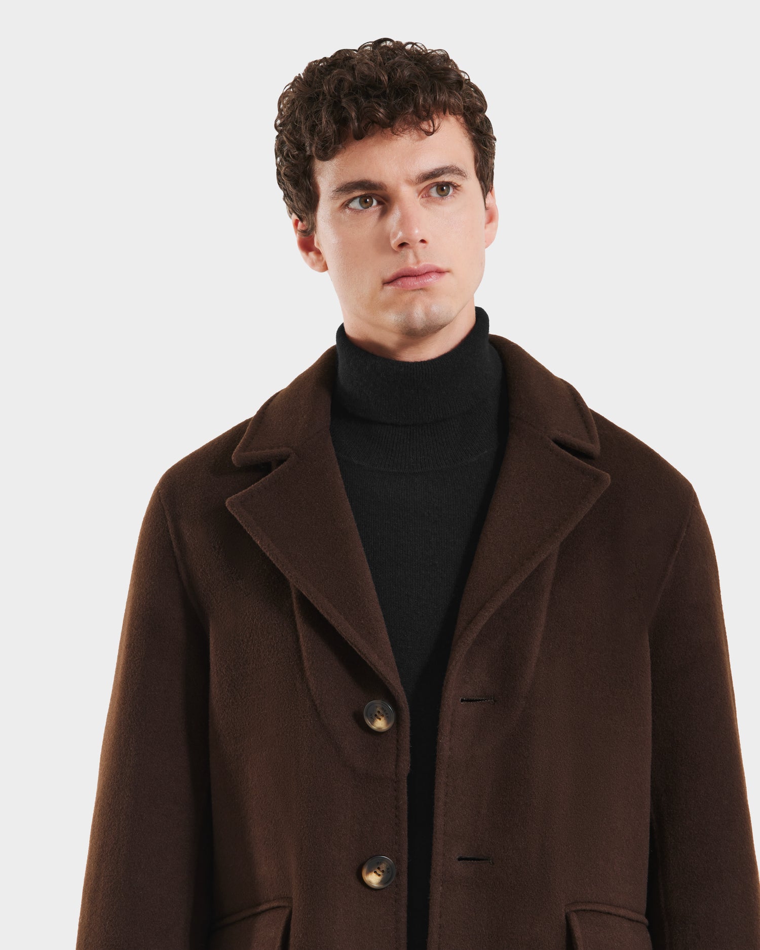 A young man with short curly hair models the Rudsak SAWYER - 6125511 Espresso mens wool blend coat over a black turtleneck, gazing to the side against a plain white background.