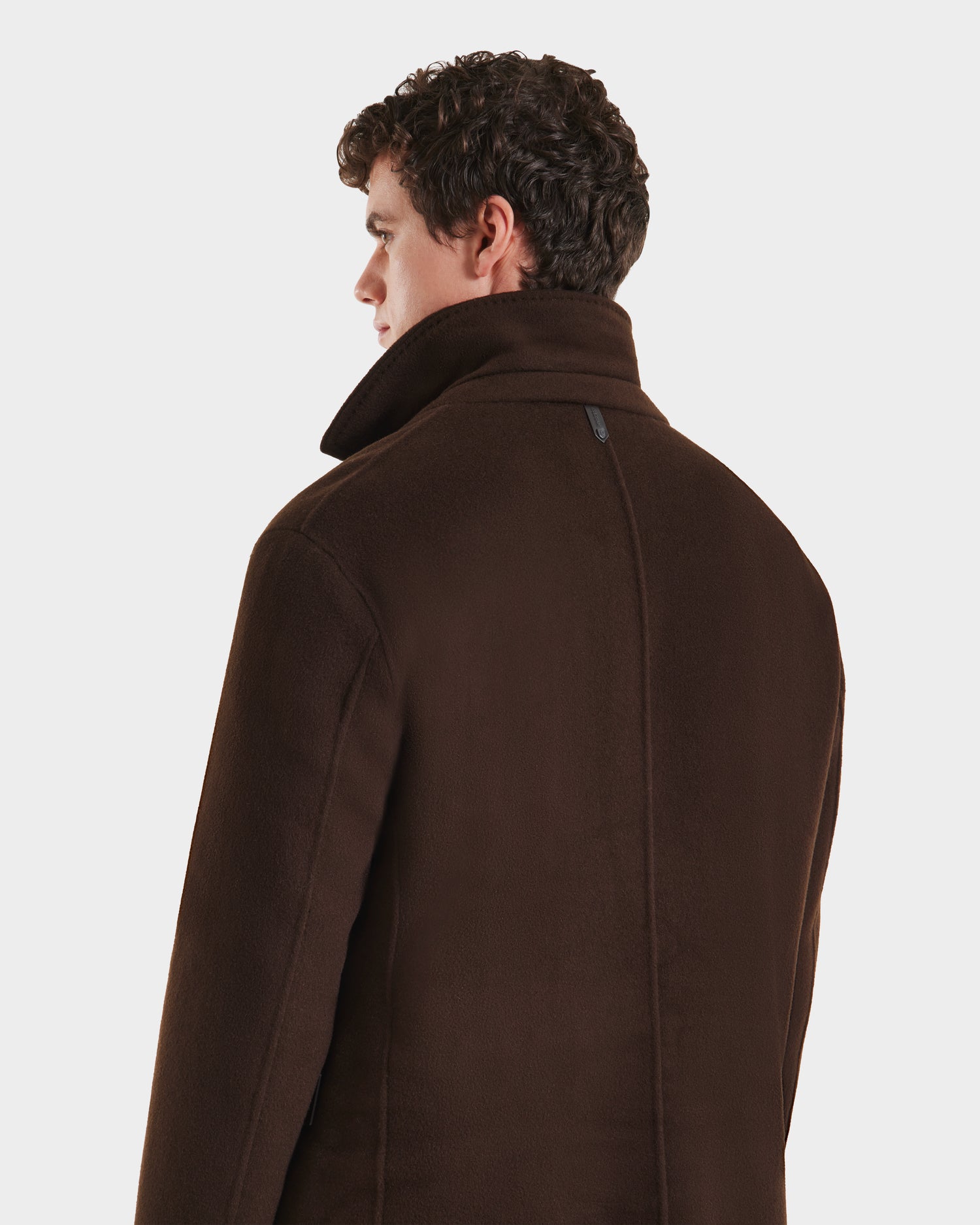 A man with curly brown hair is seen from behind wearing the Rudsak SAWYER - 6125511 Espresso men’s wool blend coat, featuring a dark brown high collar and back seam, against a plain white background.