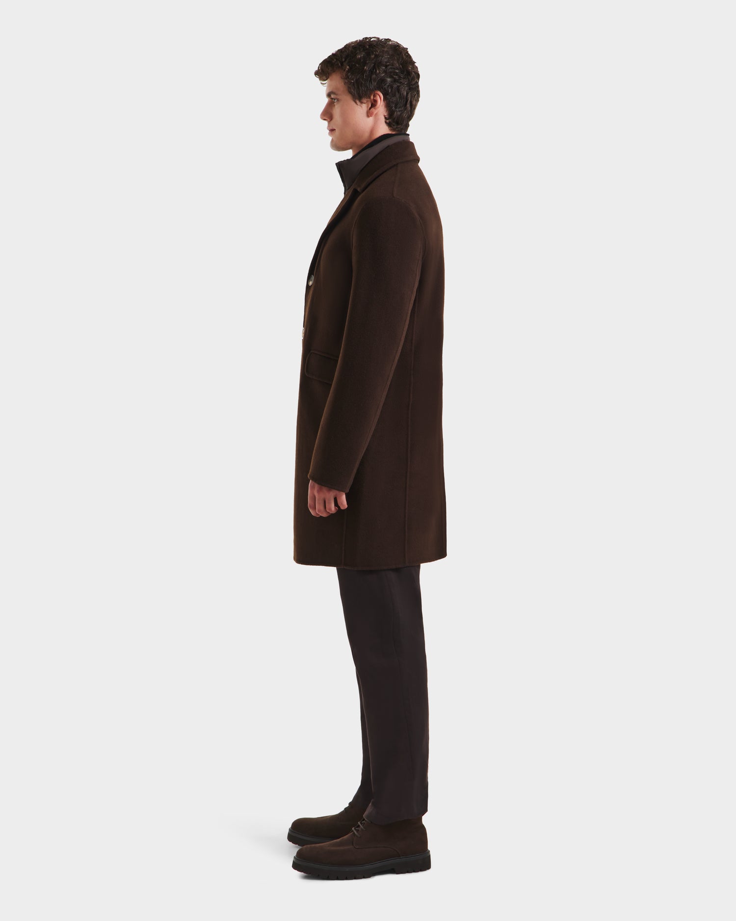 A man stands in profile facing left, wearing the Rudsak SAWYER - 6125511 Espresso men’s wool blend coat with a classic silhouette, paired with dark trousers and brown shoes against a plain white background.