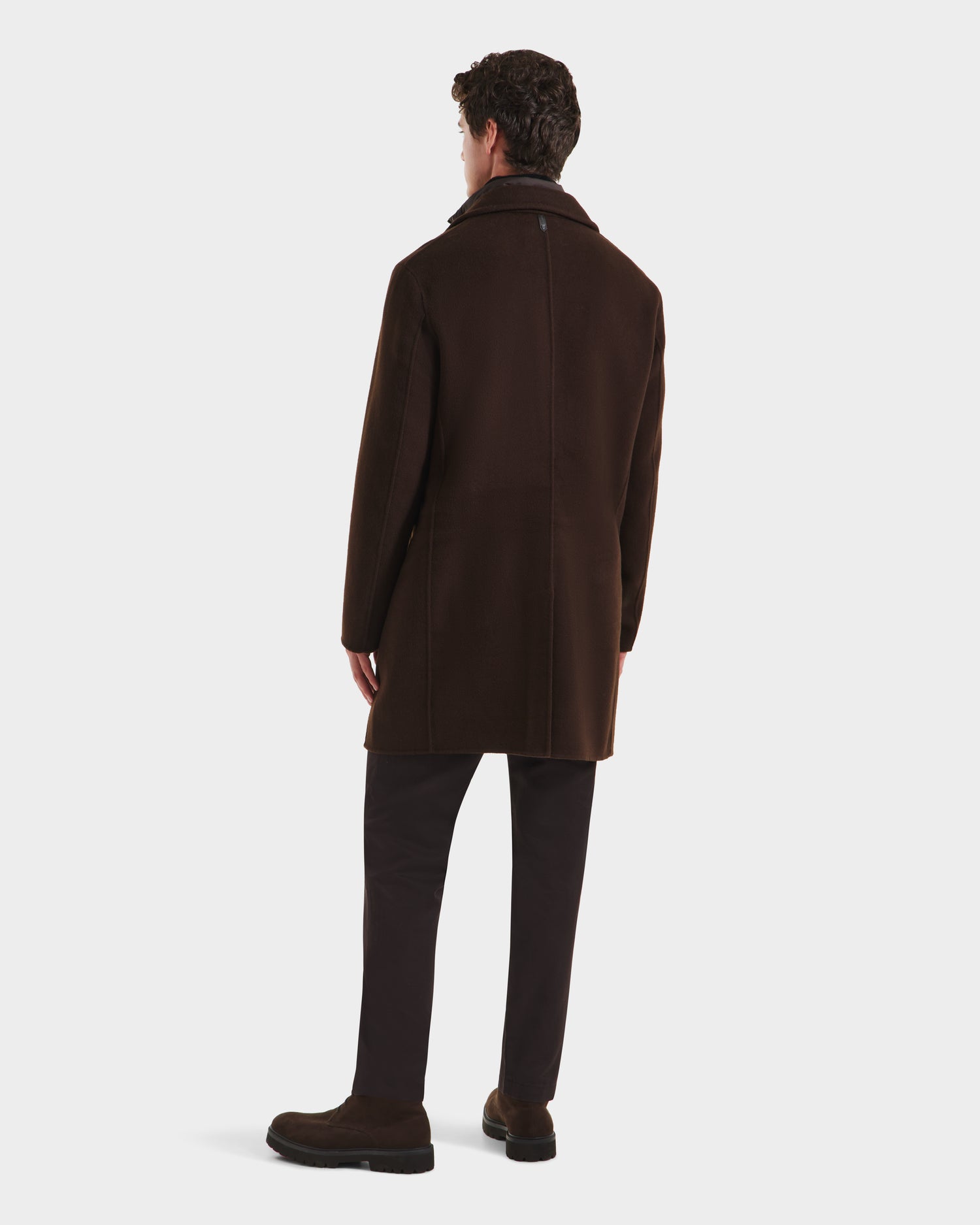 A man faces away, wearing the Rudsak SAWYER - 6125511 Espresso mens coat with a classic silhouette, paired with dark pants and shoes, set against a plain white background.