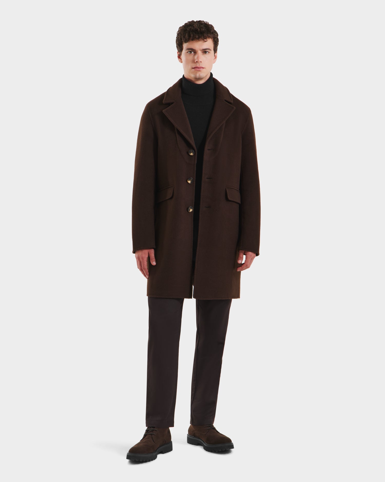 A man faces forward in a classic silhouette, wearing the Rudsak SAWYER - 6125511 Espresso mens wool blend coat with a black turtleneck, dark trousers, and brown dress shoes against a plain white background.