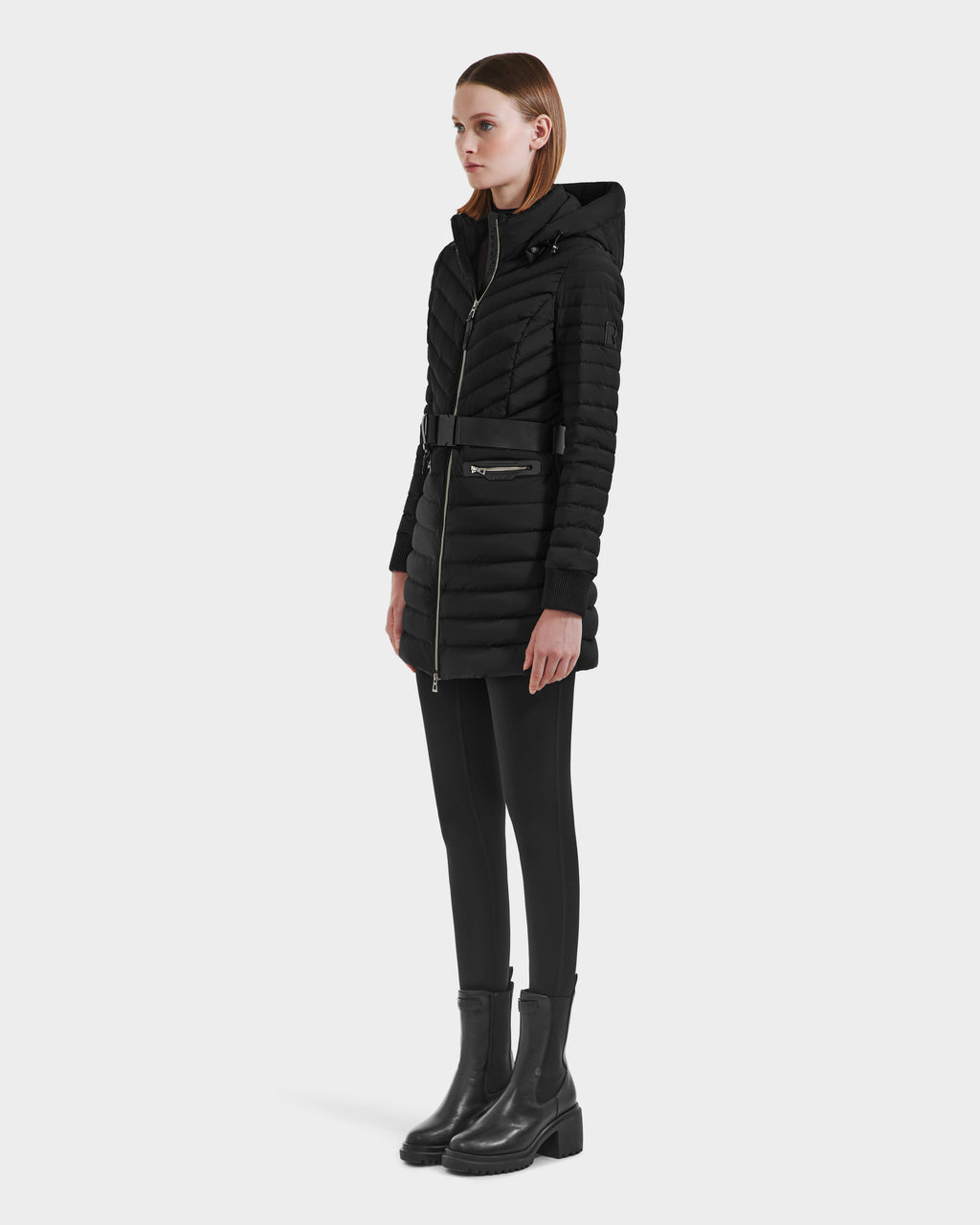 A woman faces slightly right, wearing the Rudsak SCARLETT M-M - 8125595 Black long quilted coat with a vegan leather belt, paired with black leggings and ankle boots, set against a plain white background.