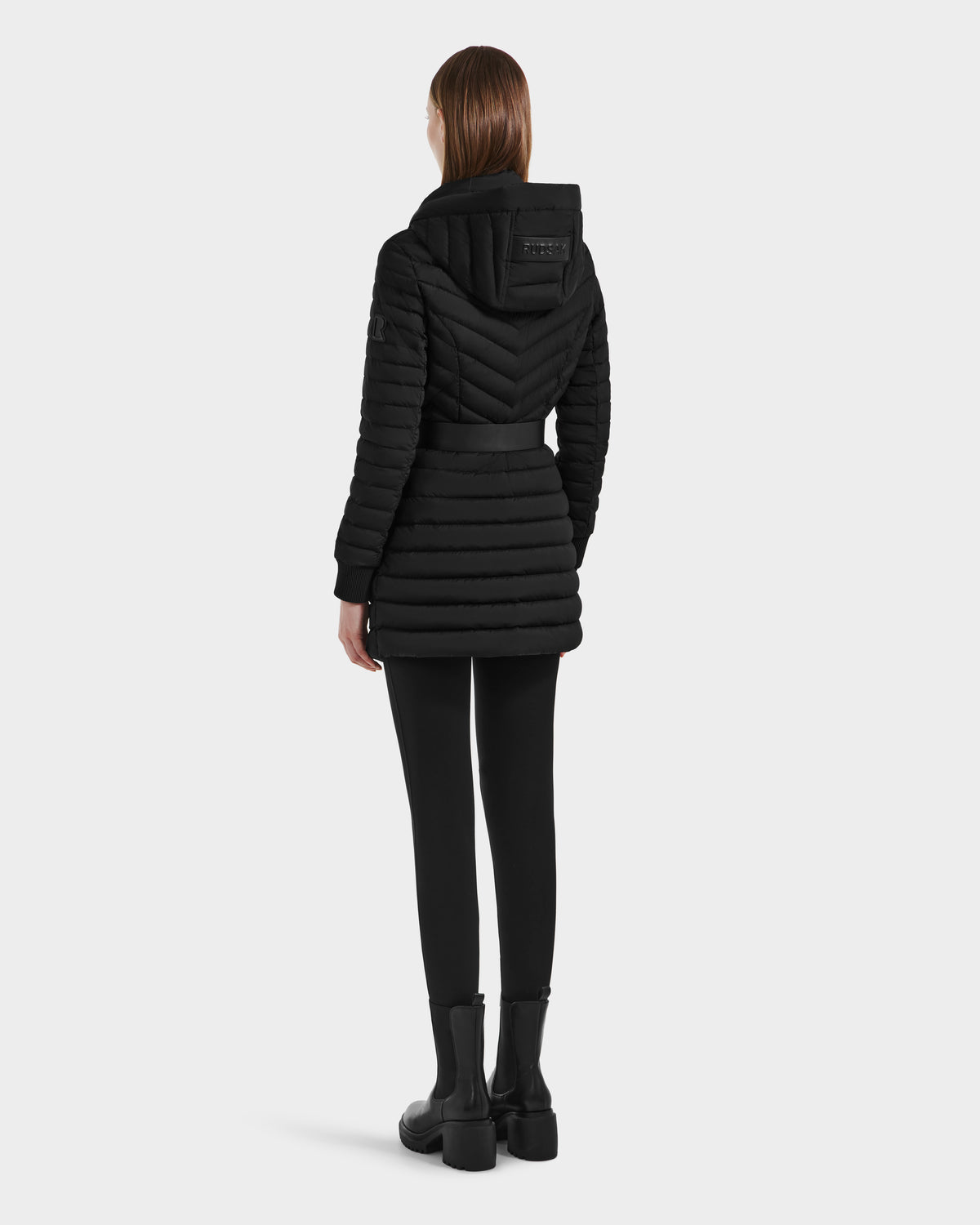 A person with straight brown hair is standing and facing away from the camera against a plain light background, wearing the Rudsak SCARLETT M-M - 8125595 Black puffer jacket with a hood, vegan leather belt, black leggings, and black boots.