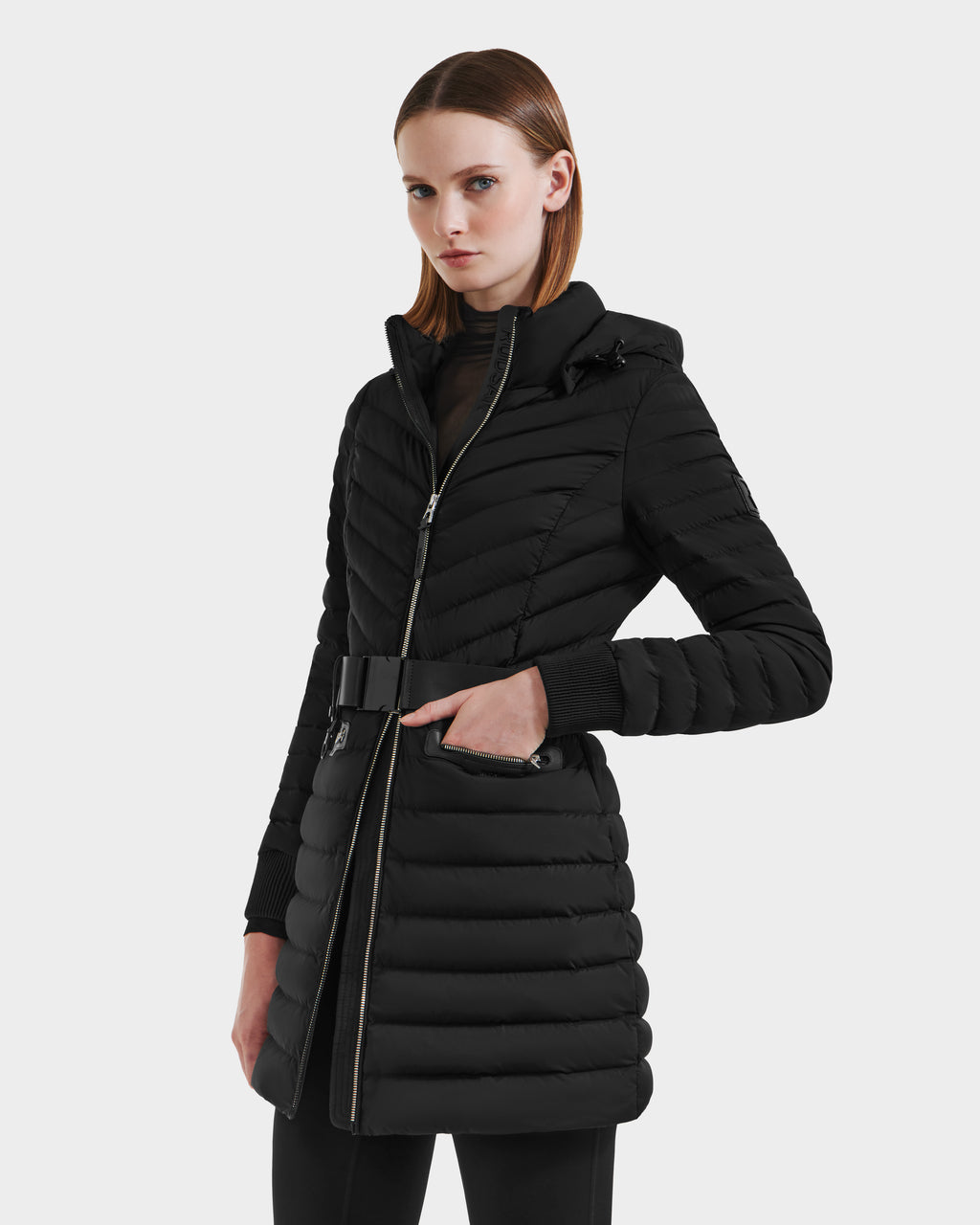 A woman wears the Rudsak SCARLETT M-M - 8125595 Black coat, featuring a quilted design, hood, vegan leather belt, and zipper, standing against a plain light gray background.