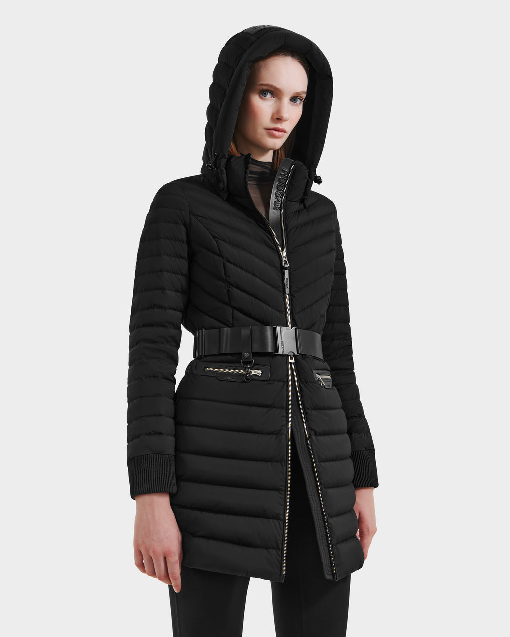 A woman stands against a plain light background wearing the Rudsak SCARLETT M-M - 8125595 Black quilted mid-length coat with a zip front, vegan leather belt, ribbed cuffs, and zippered pockets, gazing ahead with a neutral expression.
