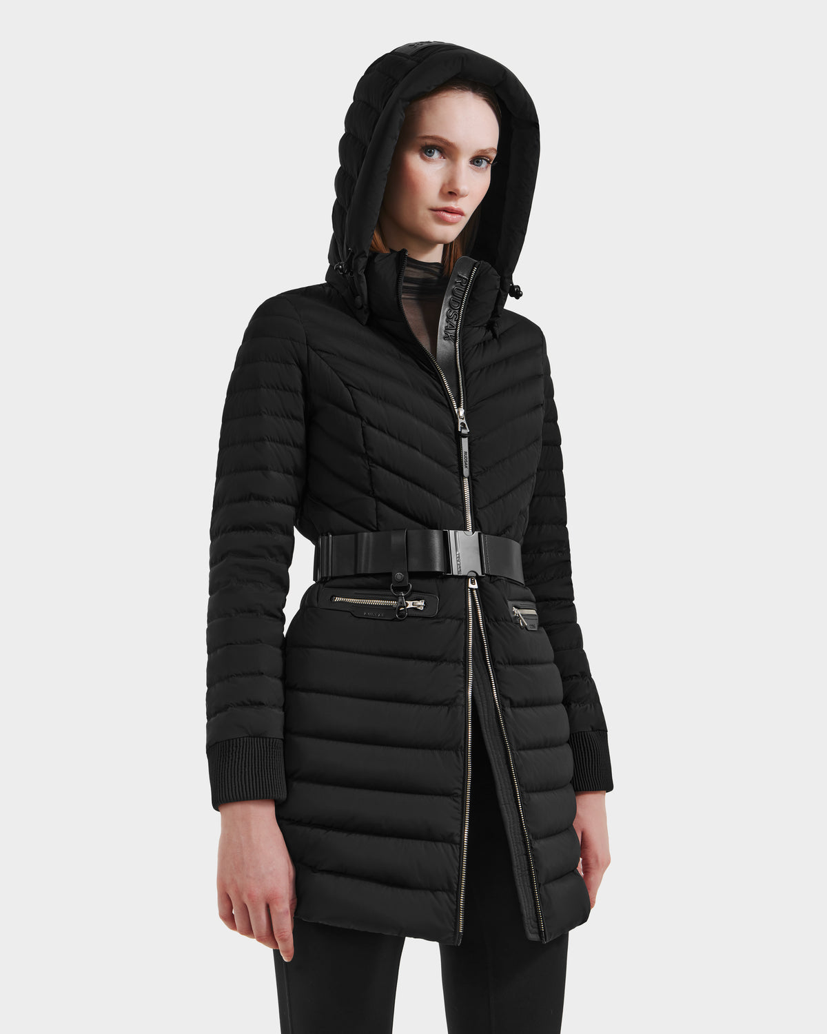 A woman stands against a plain light background wearing the Rudsak SCARLETT M-M - 8125595 Black quilted mid-length coat with a zip front, vegan leather belt, ribbed cuffs, and zippered pockets, gazing ahead with a neutral expression.