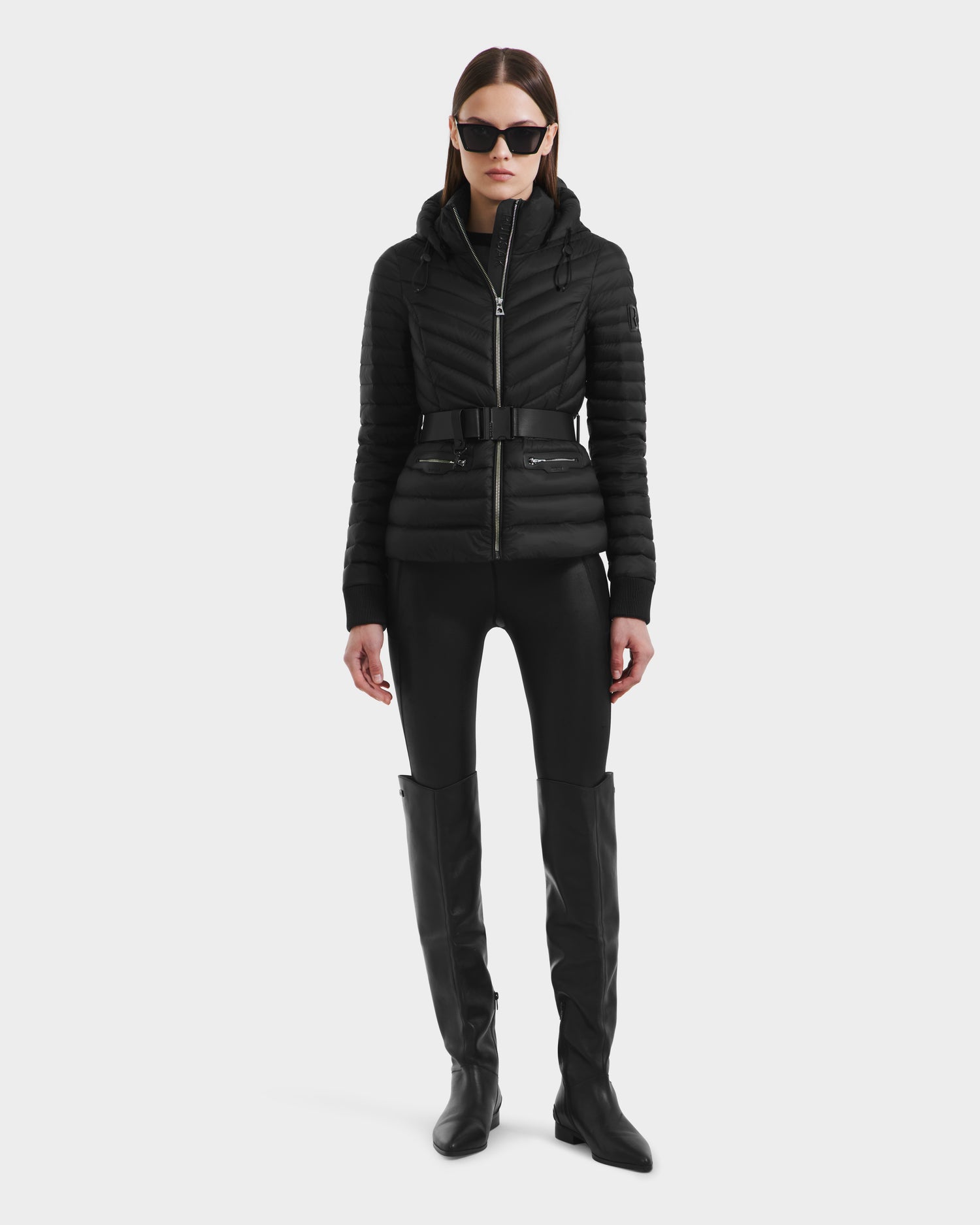 A woman faces forward in Rudsaks SCARLETT - 8125004 Black jacket featuring a vegan leather waist belt, paired with black sunglasses, fitted pants, and knee-high boots against a plain white background.