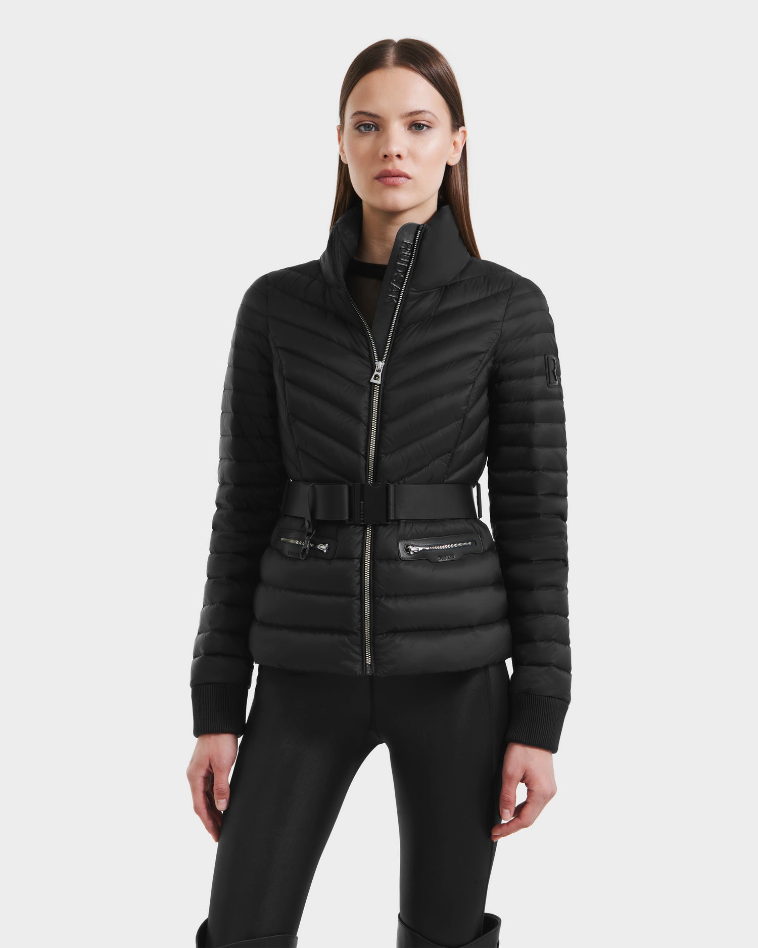 A woman faces forward in the Rudsak SCARLETT - 8125004 Black women’s lightweight down jacket with vegan leather waist belt, zippered pockets, and a high collar. The chevron quilted, wind and water-resistant coat pairs with sleek black pants on a white background.