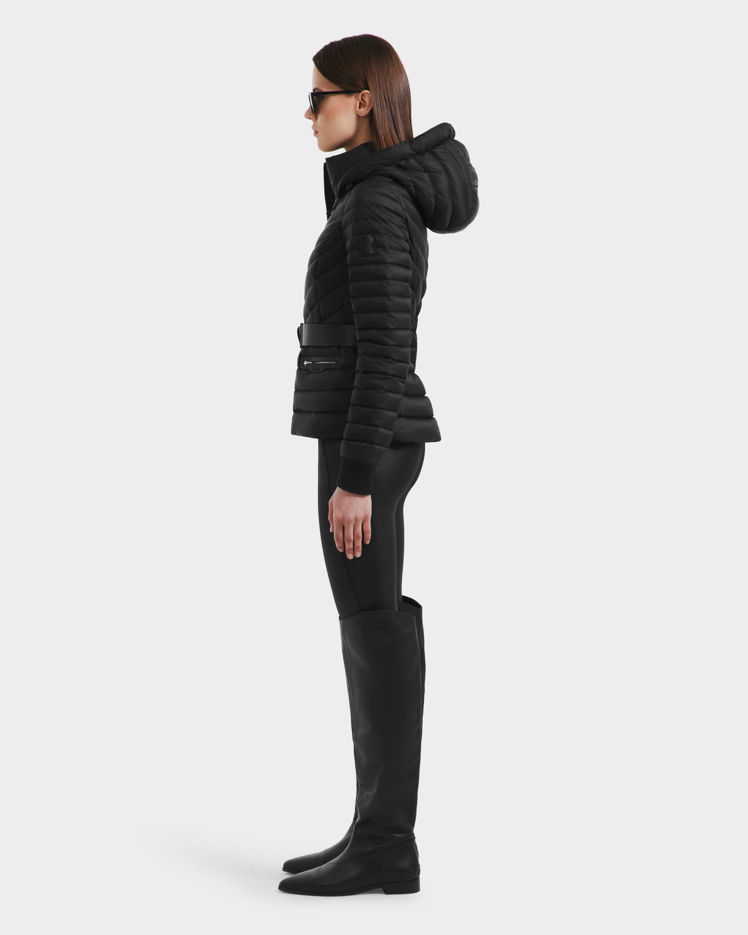 A woman in profile wears the Rudsak SCARLETT - 8125004 Black lightweight down jacket with a vegan leather waist belt, black leggings, tall black boots, and sunglasses against a plain white background.