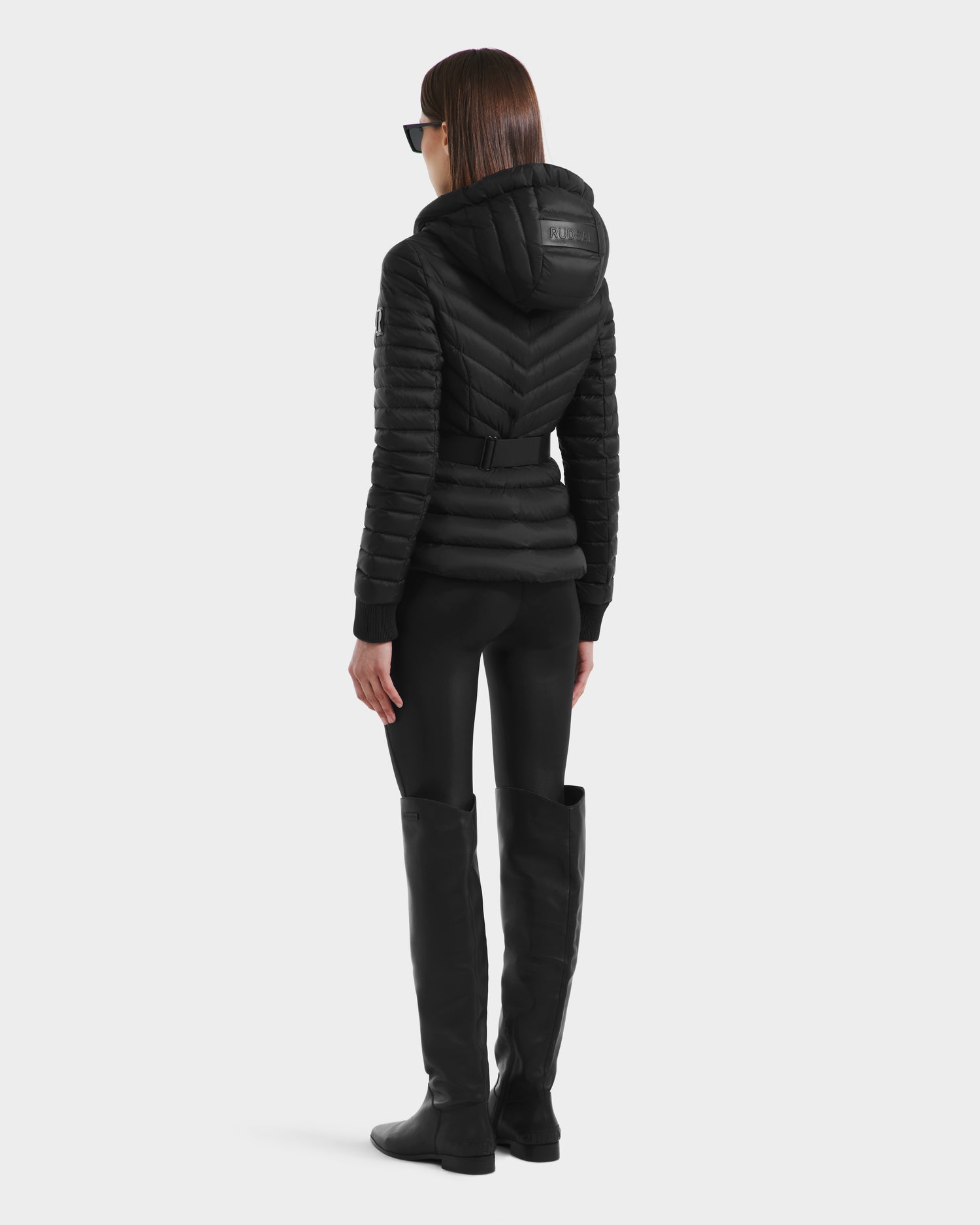 A woman with straight brown hair and black sunglasses wears the Rudsak SCARLETT - 8125004 Black fitted hooded women’s lightweight down jacket with a vegan leather waist belt, black leggings, and knee-high boots, facing away against a white background.