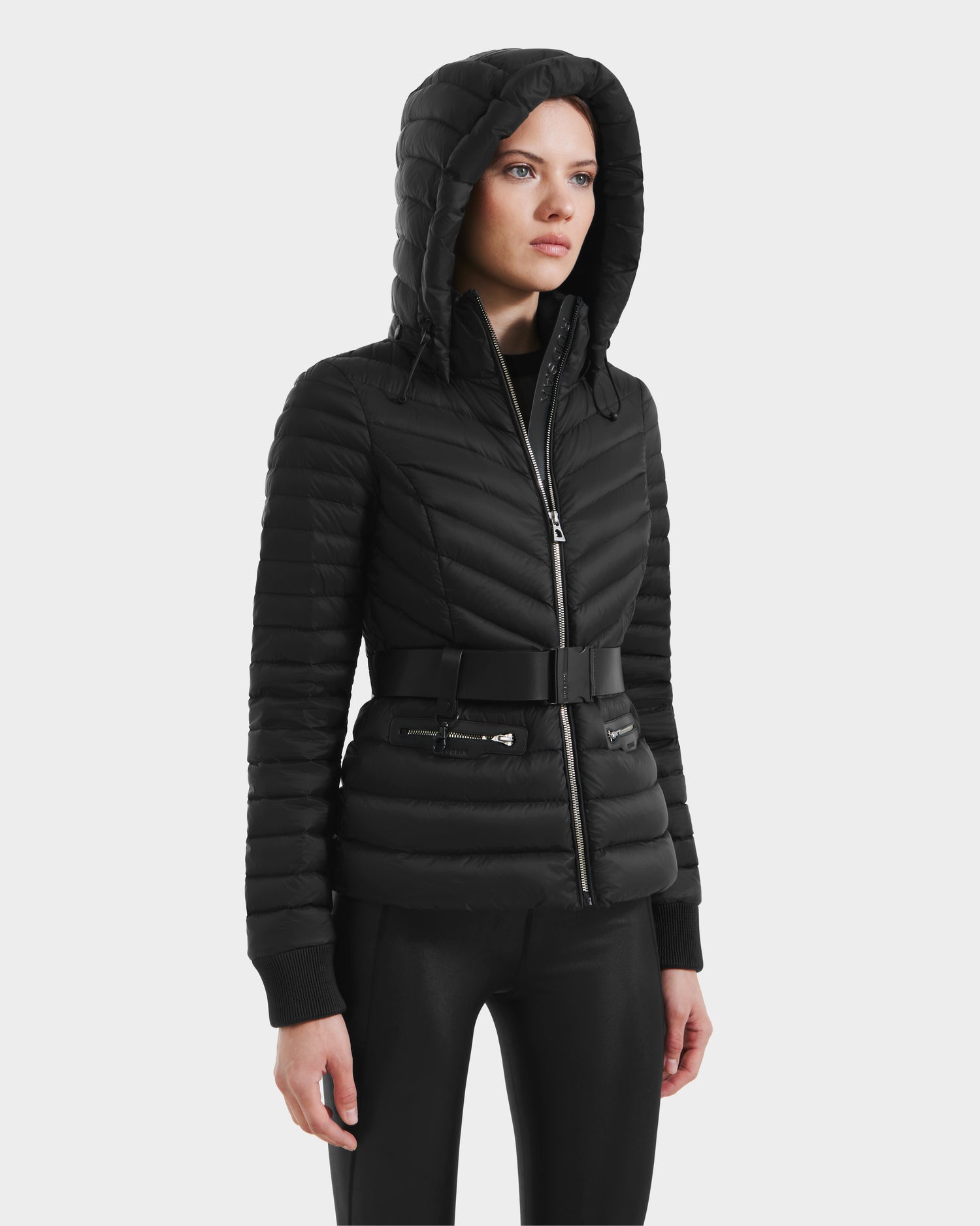 A woman models the Rudsak SCARLETT - 8125004 Black lightweight down jacket with a vegan leather waist belt, wind and water-resistant finish, zippered front and pockets, paired with black pants against a plain white background.