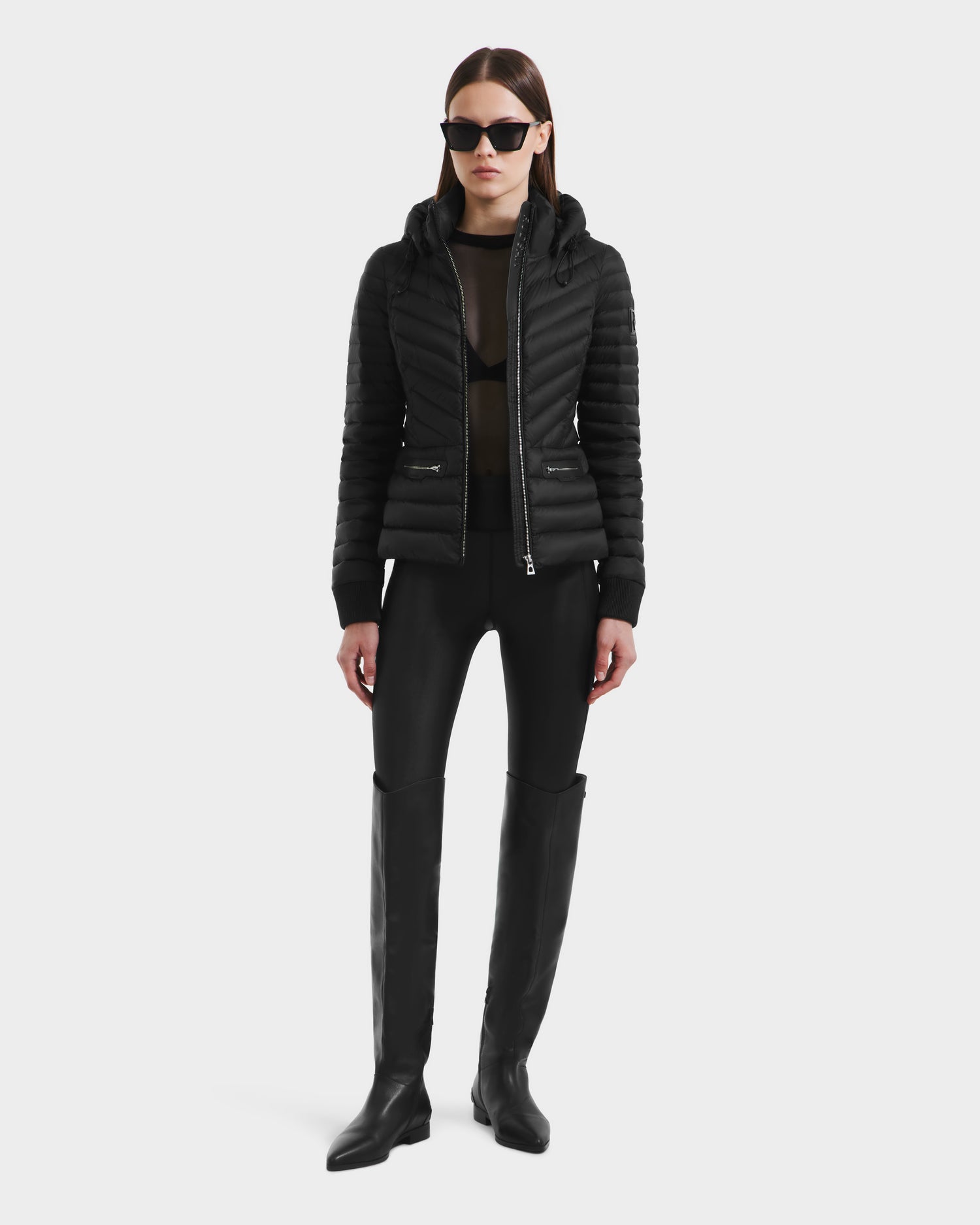A woman stands against a plain background wearing Rudsaks SCARLETT - 8125004 Black wind and water-resistant puffer jacket, paired with black sunglasses, a sheer top, fitted pants, a vegan leather waist belt, and knee-high boots.