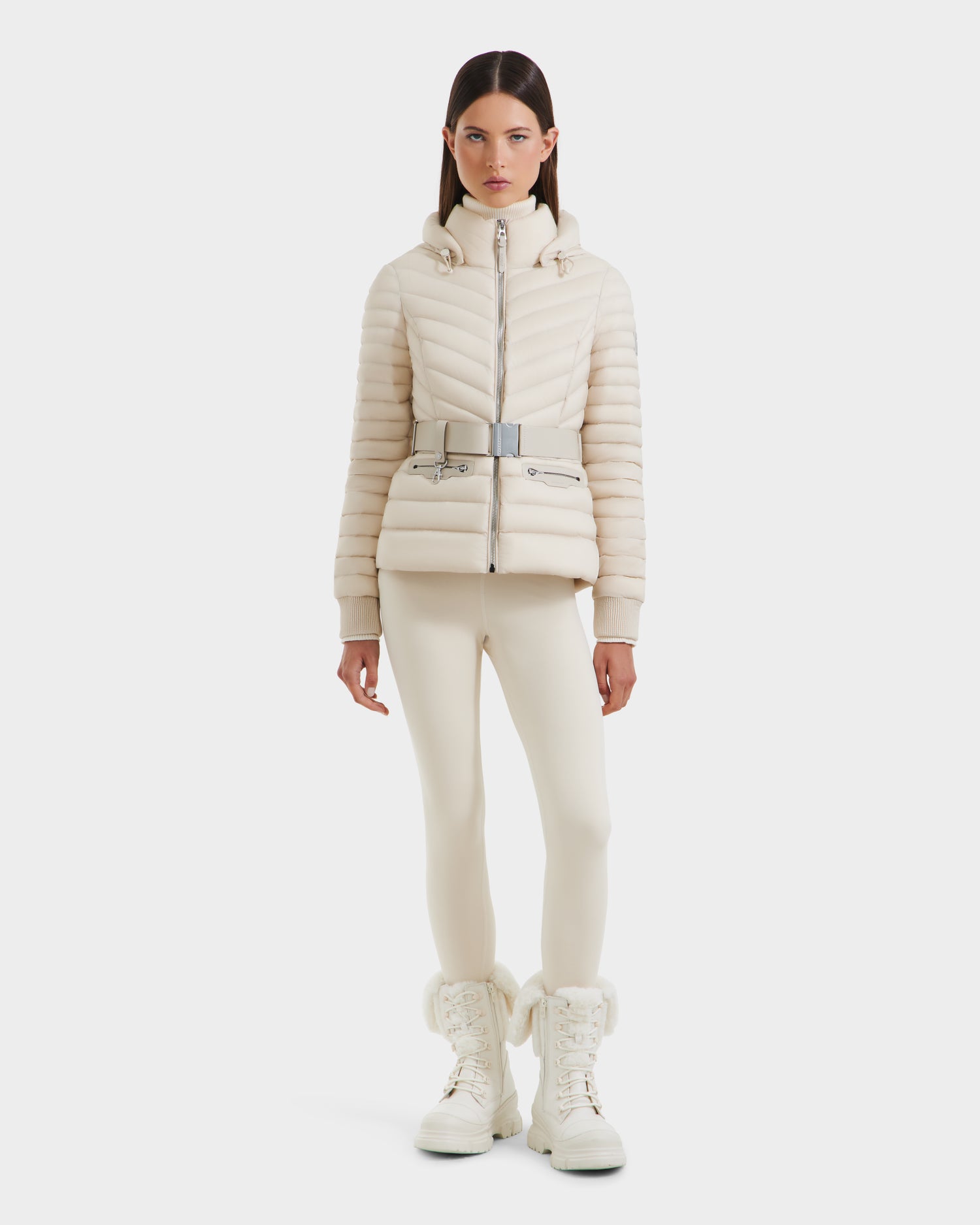 A woman faces forward, wearing the Rudsak SCARLETT - 8125004 Vanilla lightweight down jacket with a high collar, paired with fitted pants and fur-trimmed boots against a plain white background.