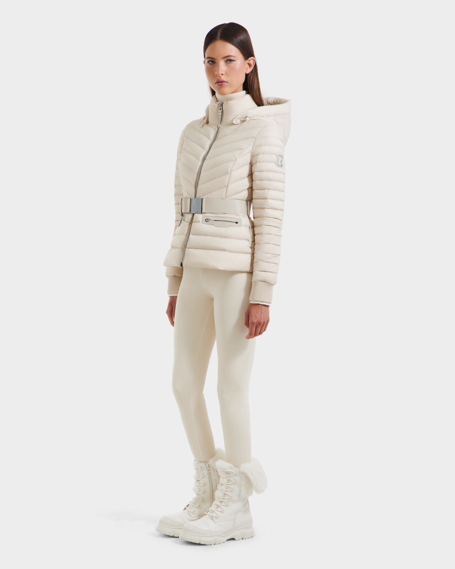 A woman with straight brown hair and a neutral expression stands against a white background, wearing the Rudsak SCARLETT - 8125004 Vanilla lightweight down jacket with a belt. She pairs it with fitted pants and fur-lined winter boots.