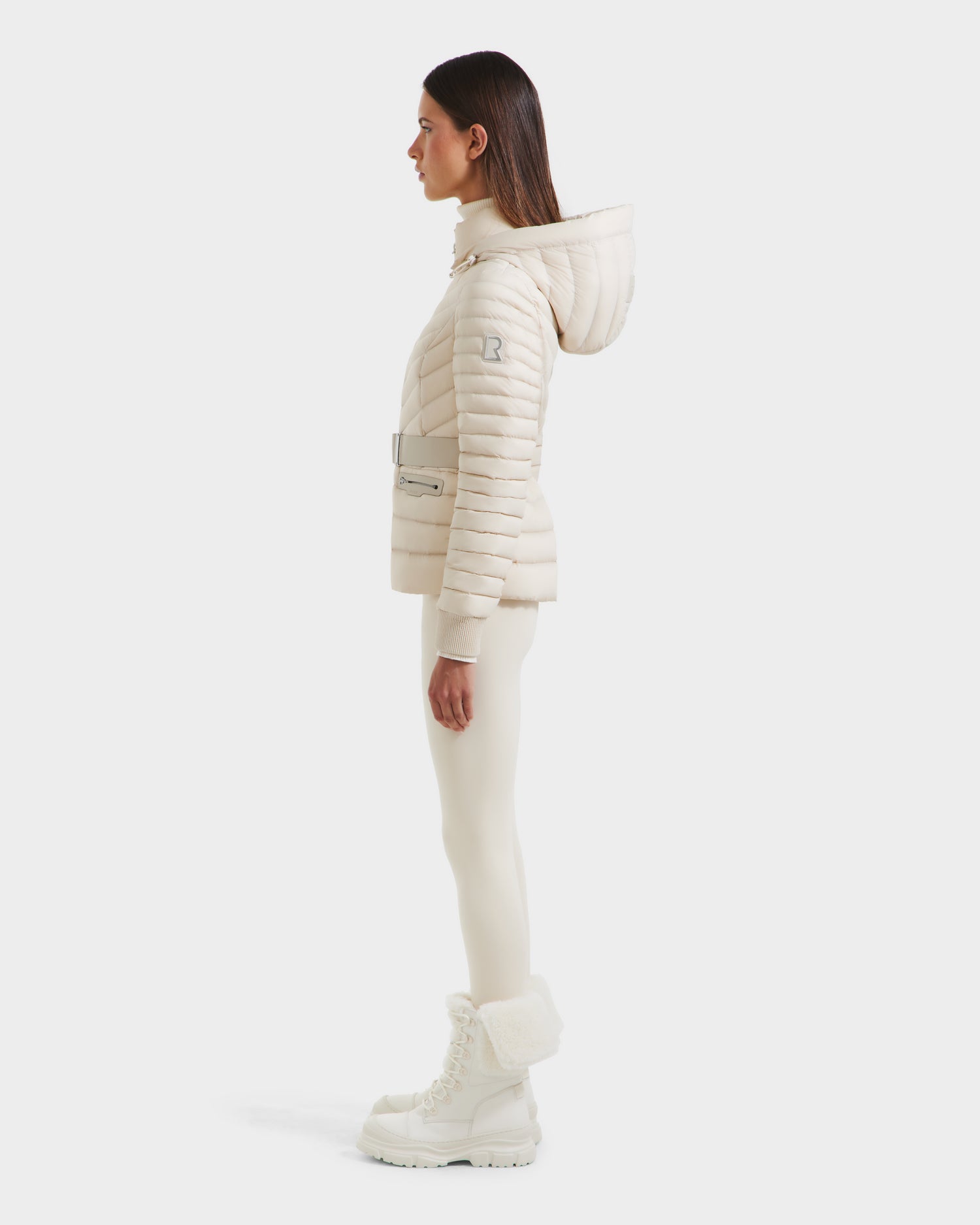 A woman stands in profile wearing the Rudsak SCARLETT - 8125004 Vanilla lightweight down jacket with a hood, matching leggings, and white winter boots, photographed against a plain white background.