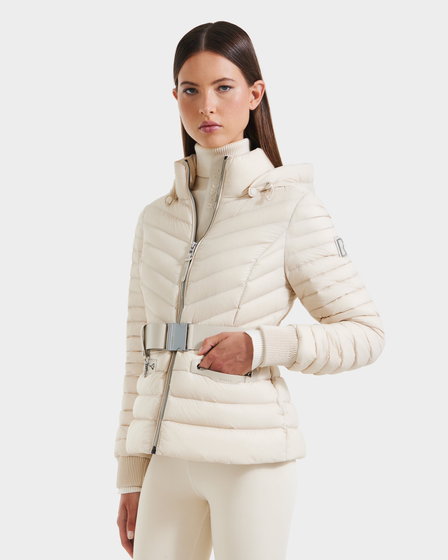 A woman wears the Rudsak SCARLETT - 8125004 Vanilla lightweight down jacket with a high collar and zipper, paired with matching cream pants, as she stands against a plain white background.