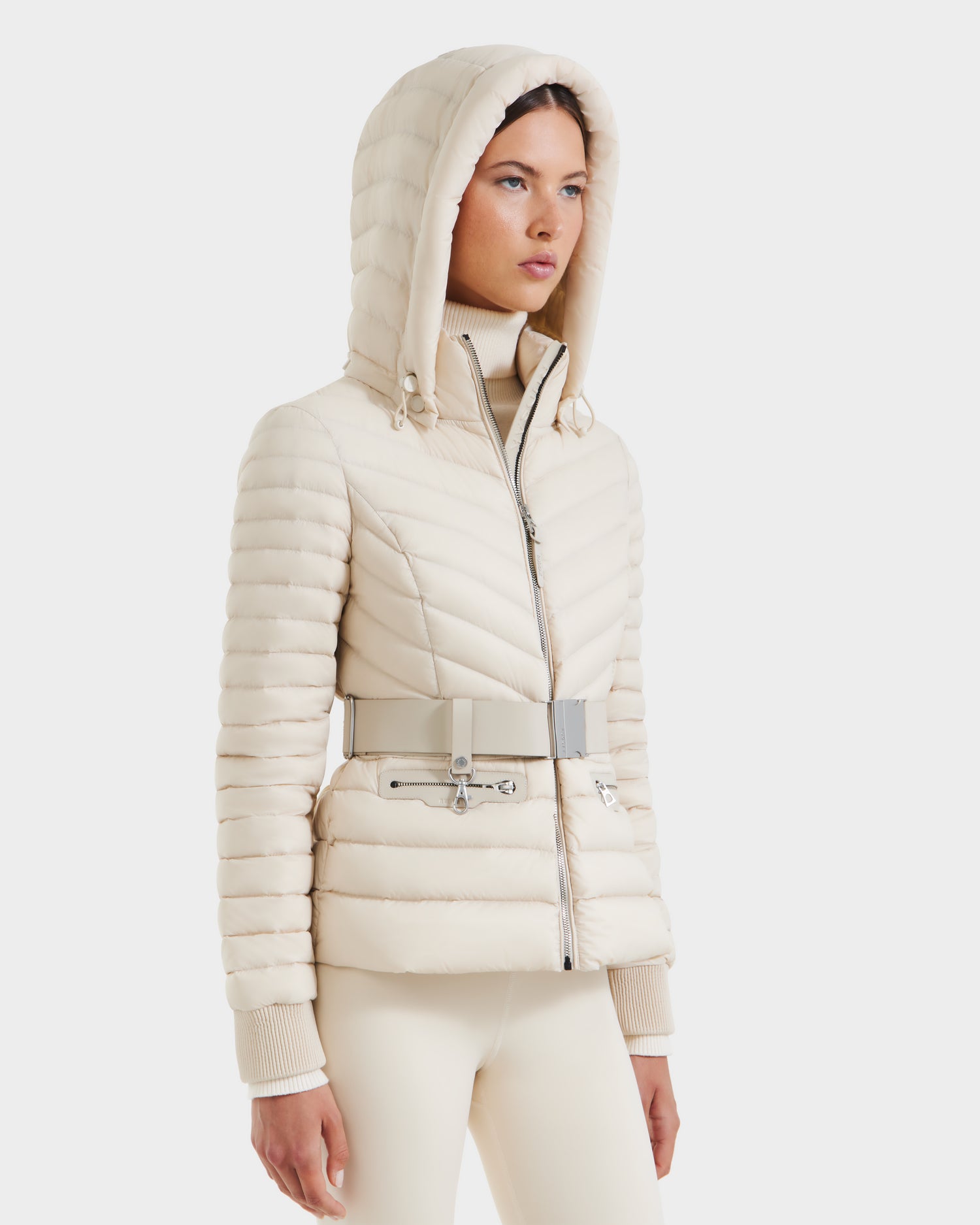 A woman wears the Rudsak SCARLETT - 8125004 Vanilla, a stylish cream-colored quilted women’s lightweight down jacket with a wide belt and zipper, paired with matching pants, standing against a white background.