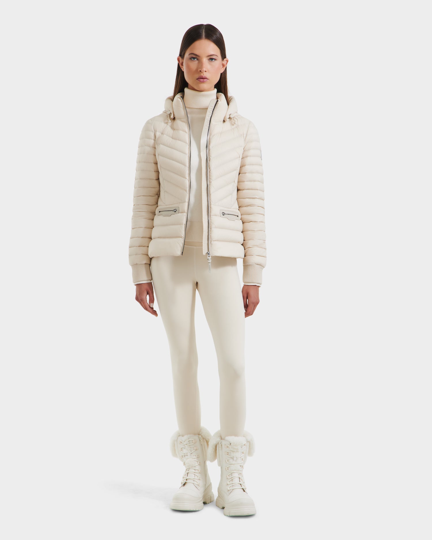 A woman faces forward against a white background, wearing the Rudsak SCARLETT - 8125004 Vanilla lightweight down jacket, paired with fitted pants, a turtleneck sweater, and fur-trimmed boots.