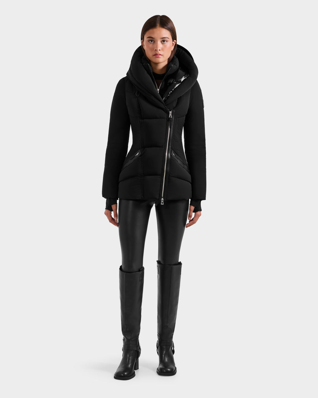 A person stands facing forward, wearing the fitted silhouette Rudsak SHALI - 8124588 Black winter jacket, black leather-look leggings, and knee-high black boots against a plain light gray background.