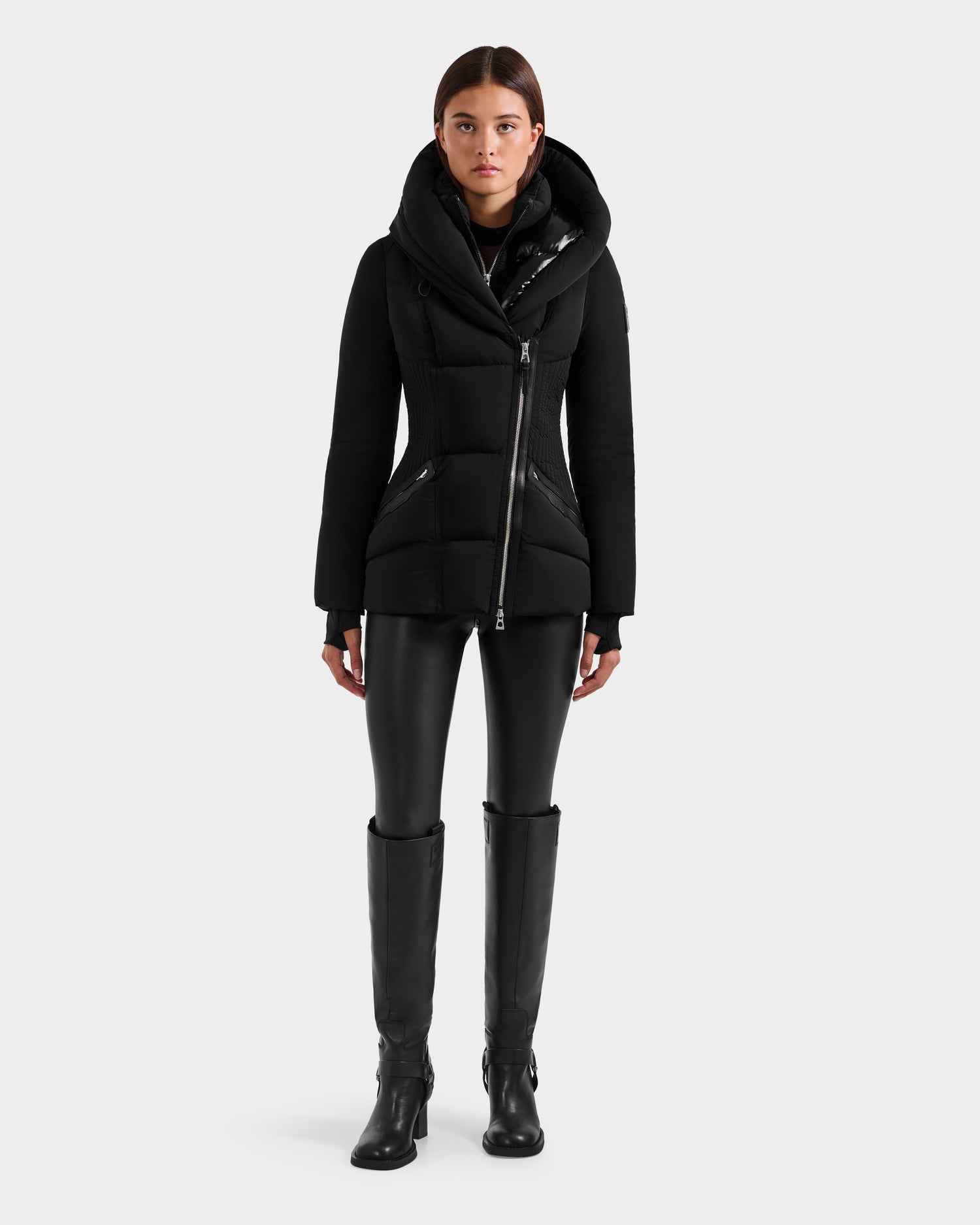 A person stands facing forward, wearing the fitted silhouette Rudsak SHALI - 8124588 Black winter jacket, black leather-look leggings, and knee-high black boots against a plain light gray background.